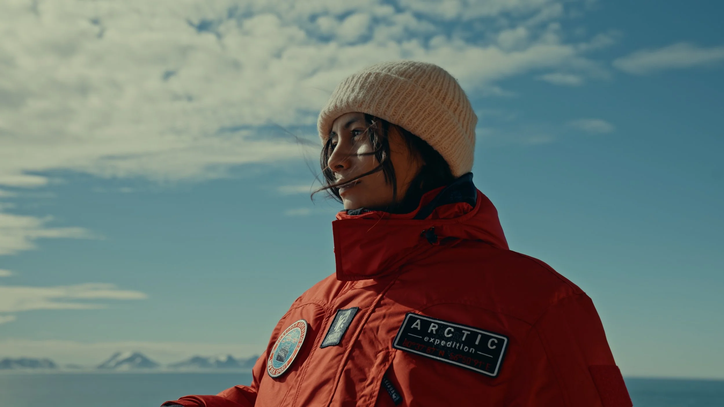 A woman in a red expedition jacket with an 'ARCTIC' patch and a beige knit beanie looks contemplatively to the side against a cloudy sky and distant mountains over water.