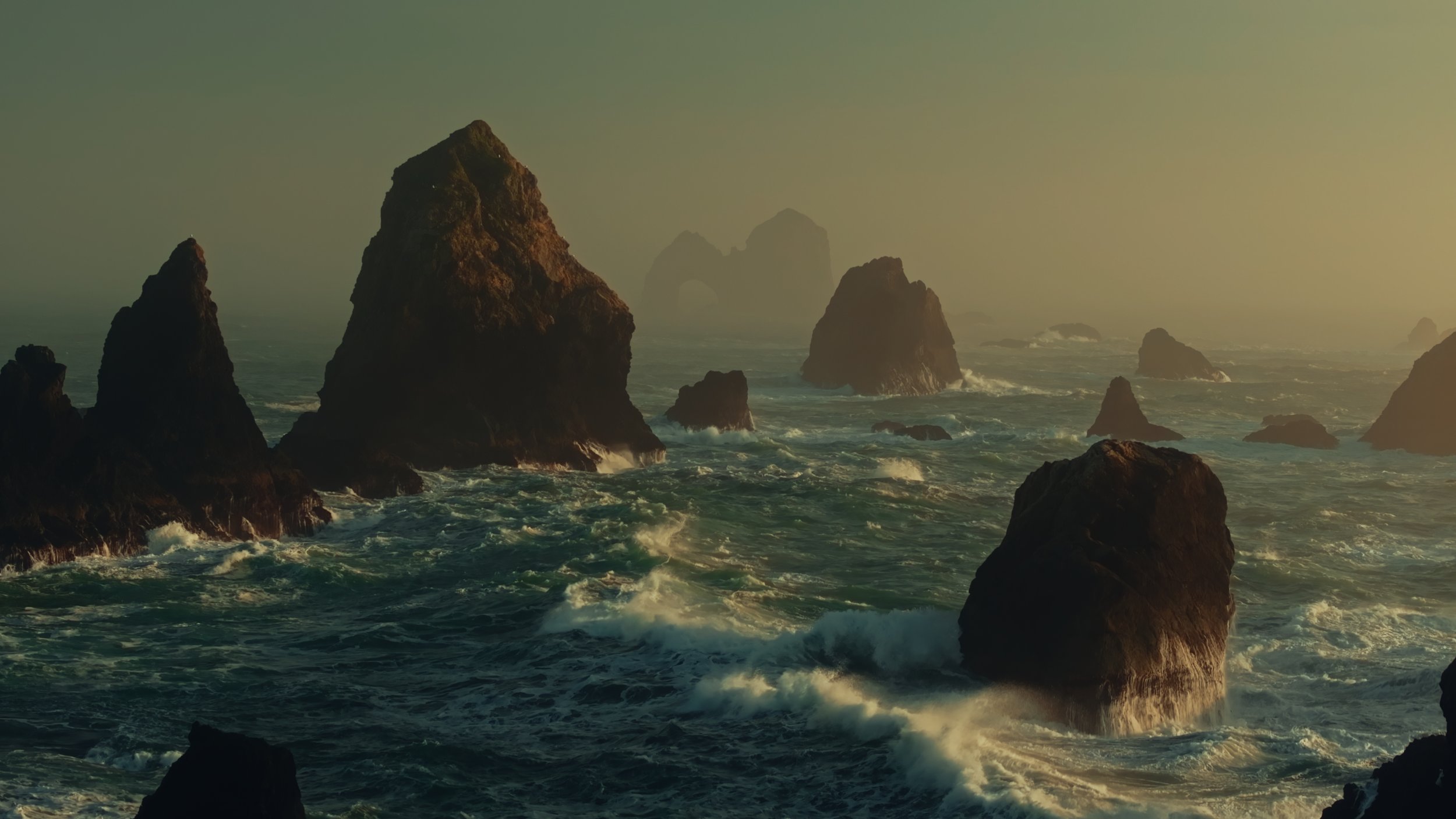 View of rugged sea stacks in ocean with waves crashing, during sunset or sunrise with hazy sky in the background.