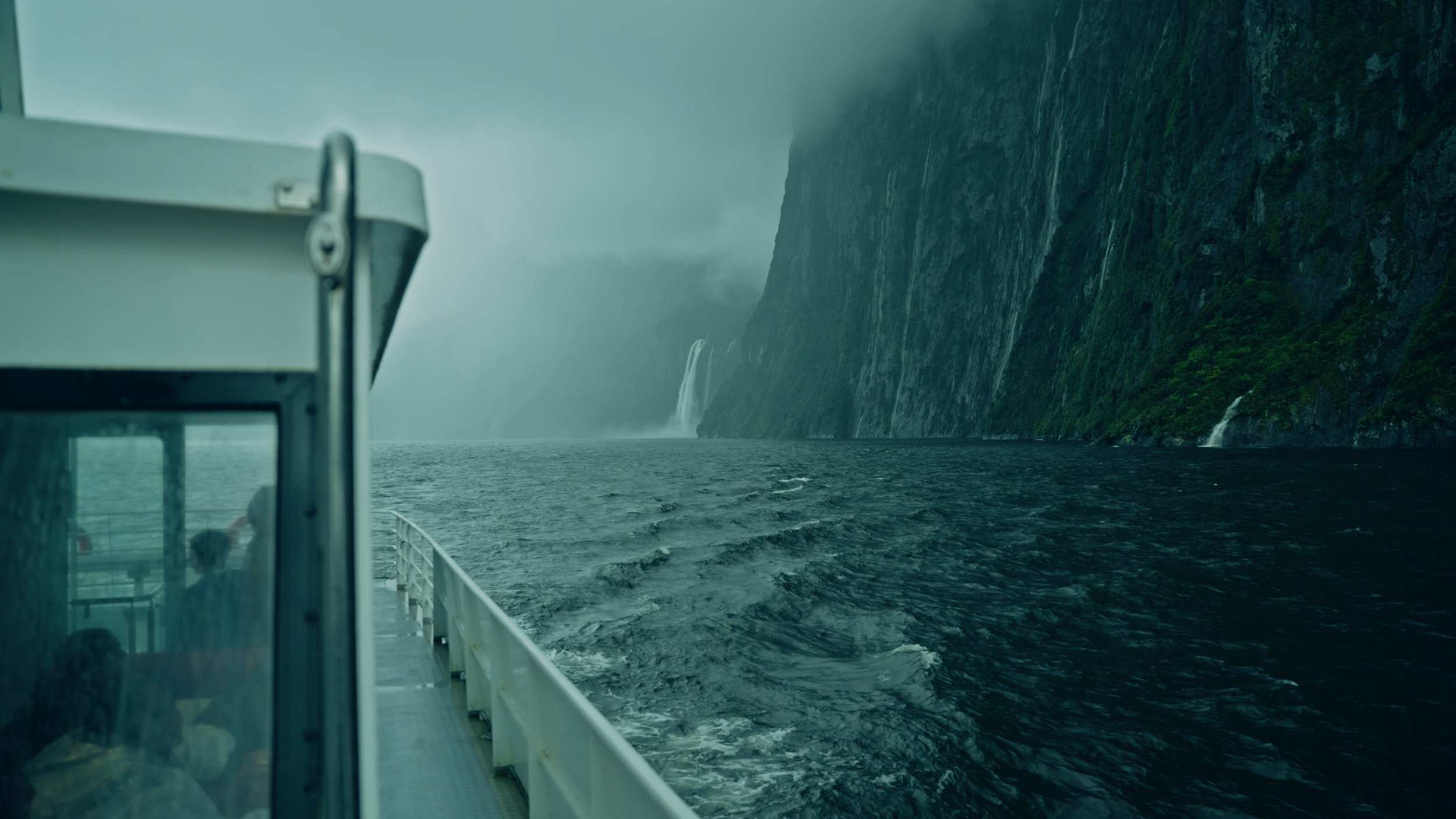 View from a boat on the water near steep, green-covered cliffs with waterfalls in the distance, possibly in a fjord.