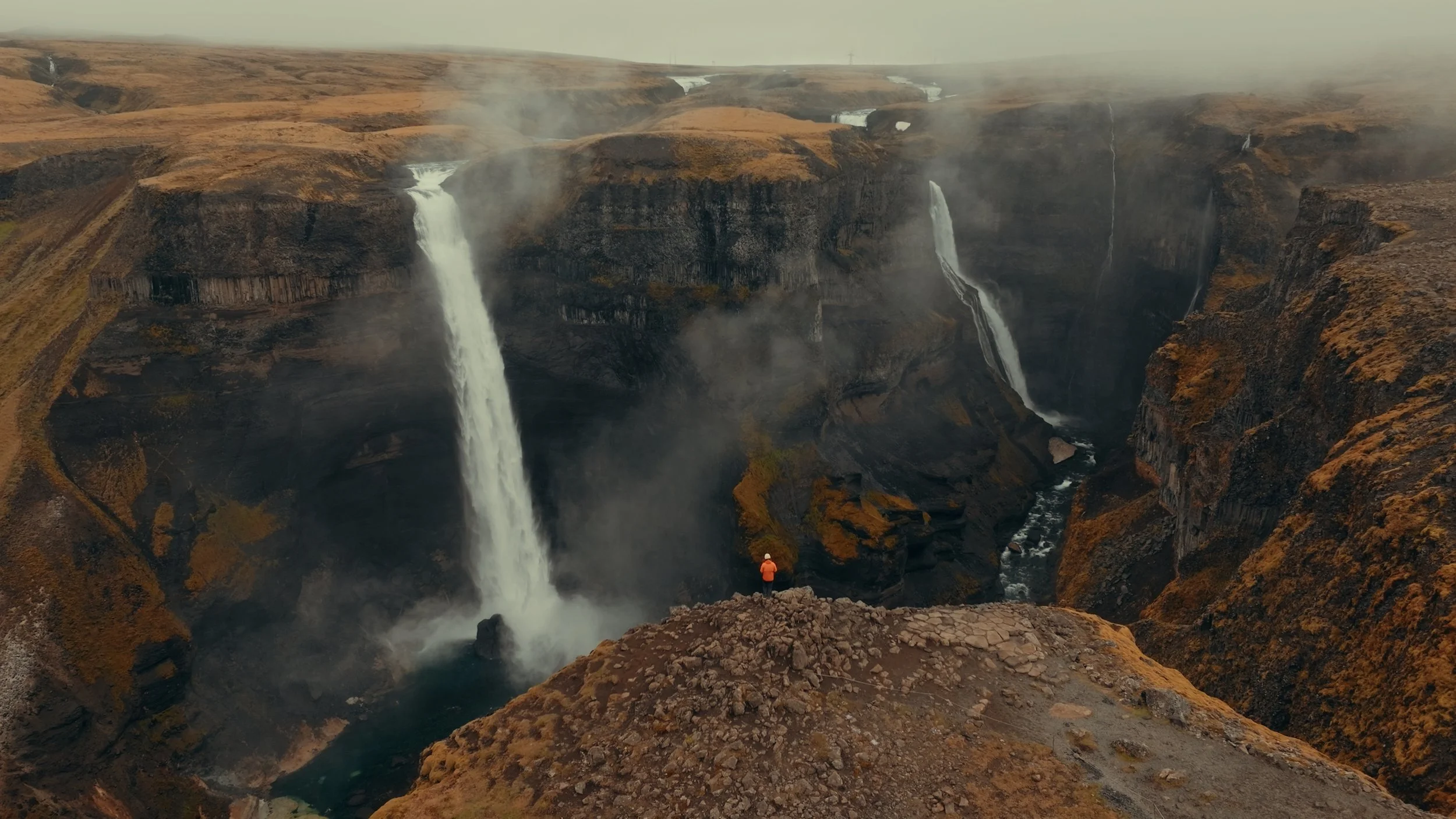 A person in an orange jacket and white hat standing on rocky ground overlooking a large canyon with three waterfalls and mist rising from the water below.