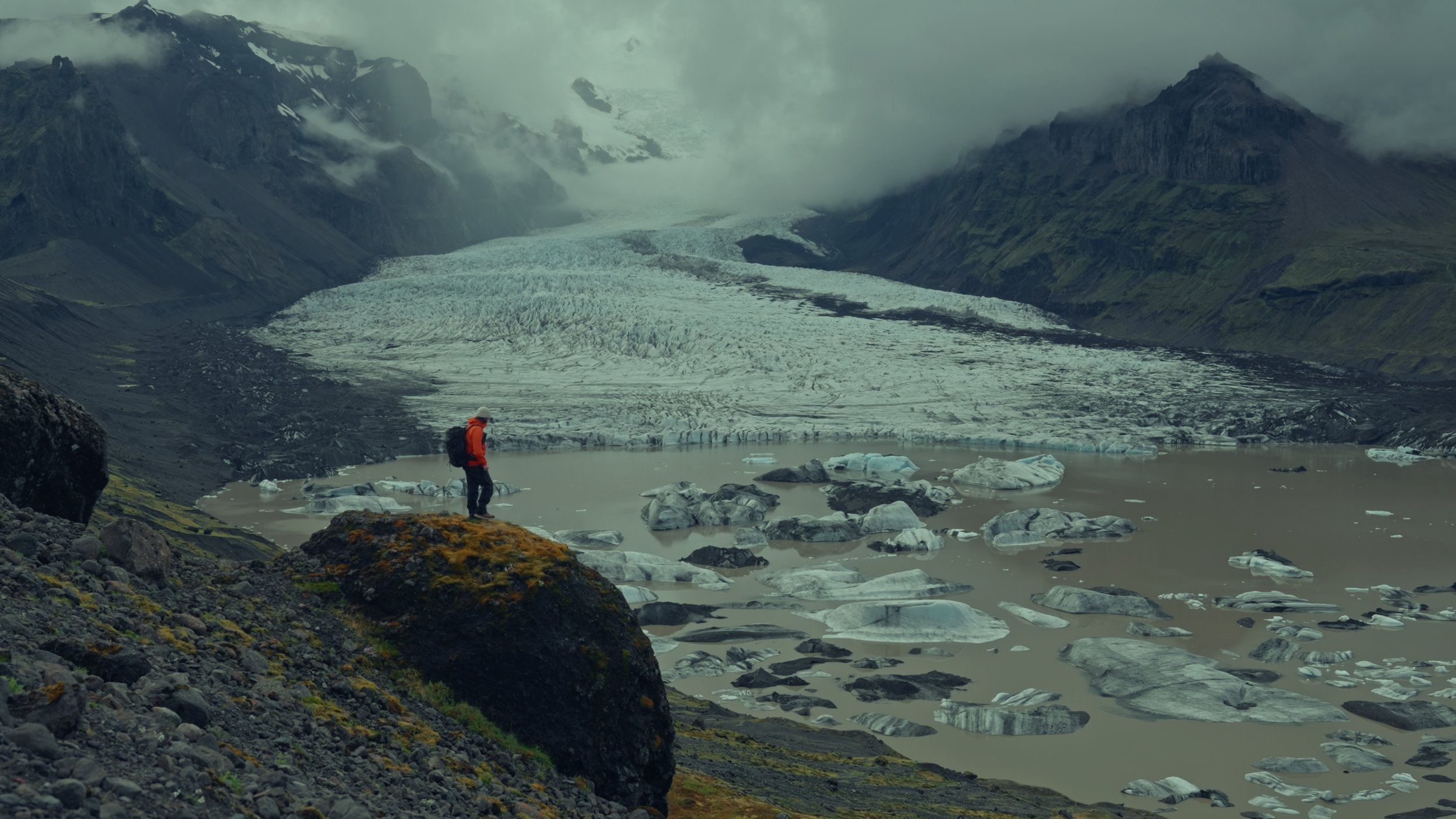 A person in an orange jacket and backpack standing on rocks near a glacier with ice chunks and a mountain range in the background.