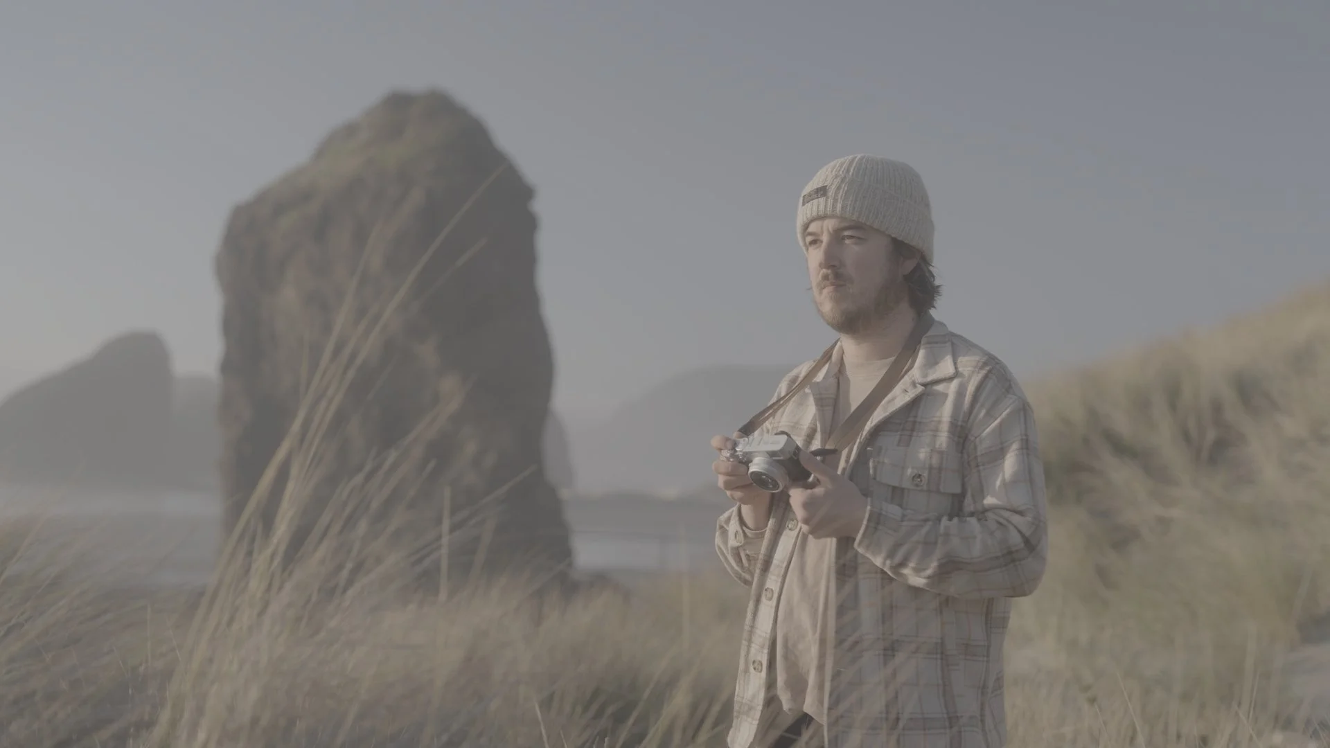 A man wearing a beige beanie and plaid shirt holding a camera, standing outdoors in a grassy area with rock formations in the background.