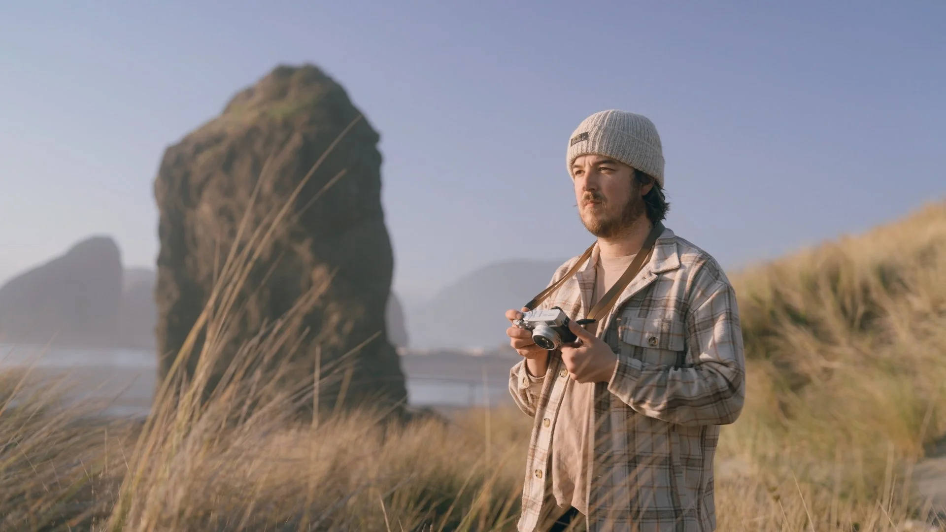 Man on beach holding camera, wearing beanie and checkered shirt, with large rock formation in background