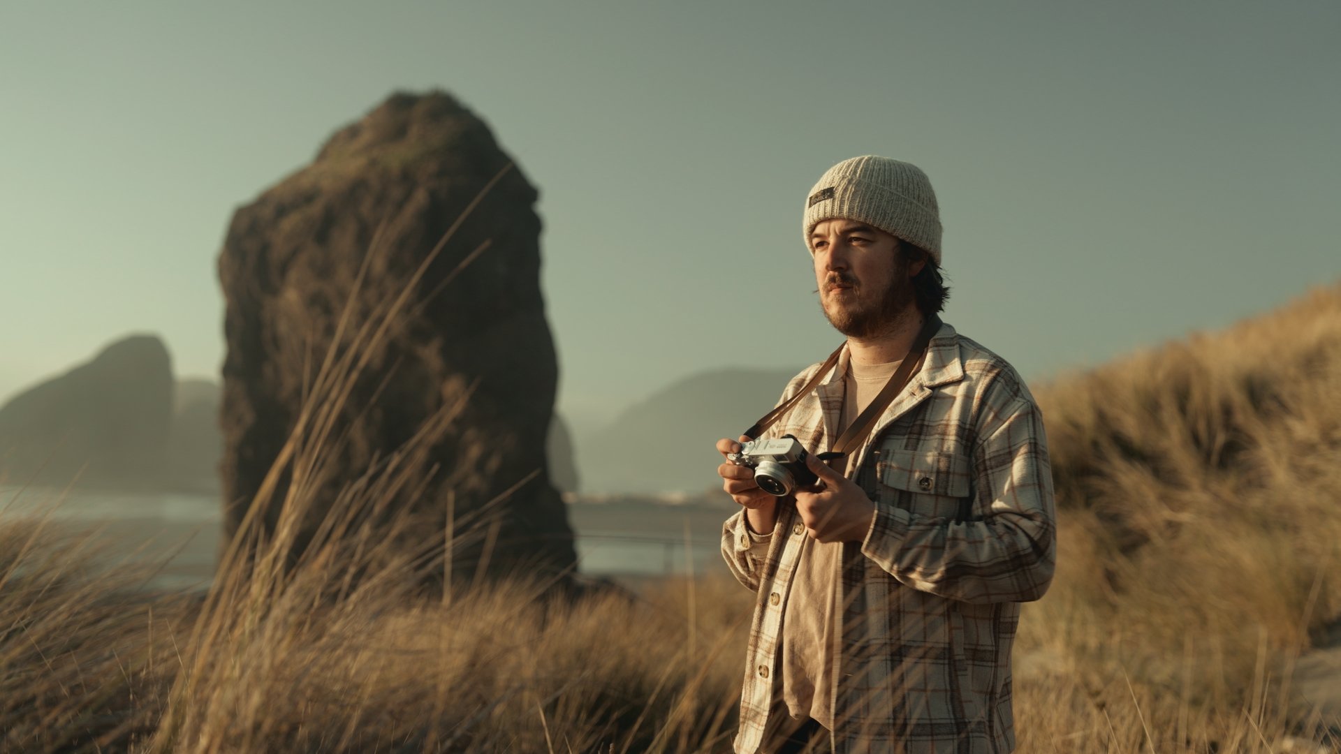A man with a camera around his neck standing outdoors during what appears to be late afternoon or early evening in a grassy coastal area with large rock formations in the background.