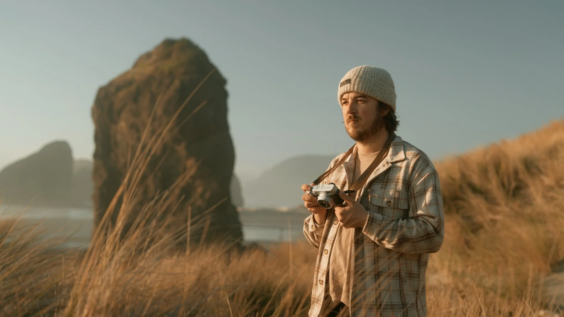 A man wearing a beige plaid shirt and a gray knit beanie is holding a camera and standing outdoors in a grassy area with large rock formations in the background during sunset.