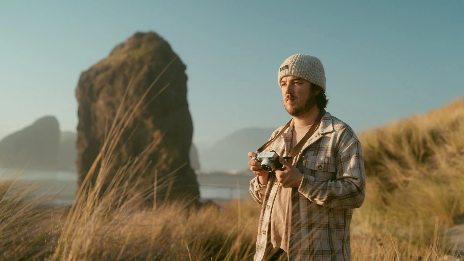 A man wearing a gray knit hat and plaid shirt standing outdoors, holding a camera, with large rock formations and grassy dunes in the background during sunset.