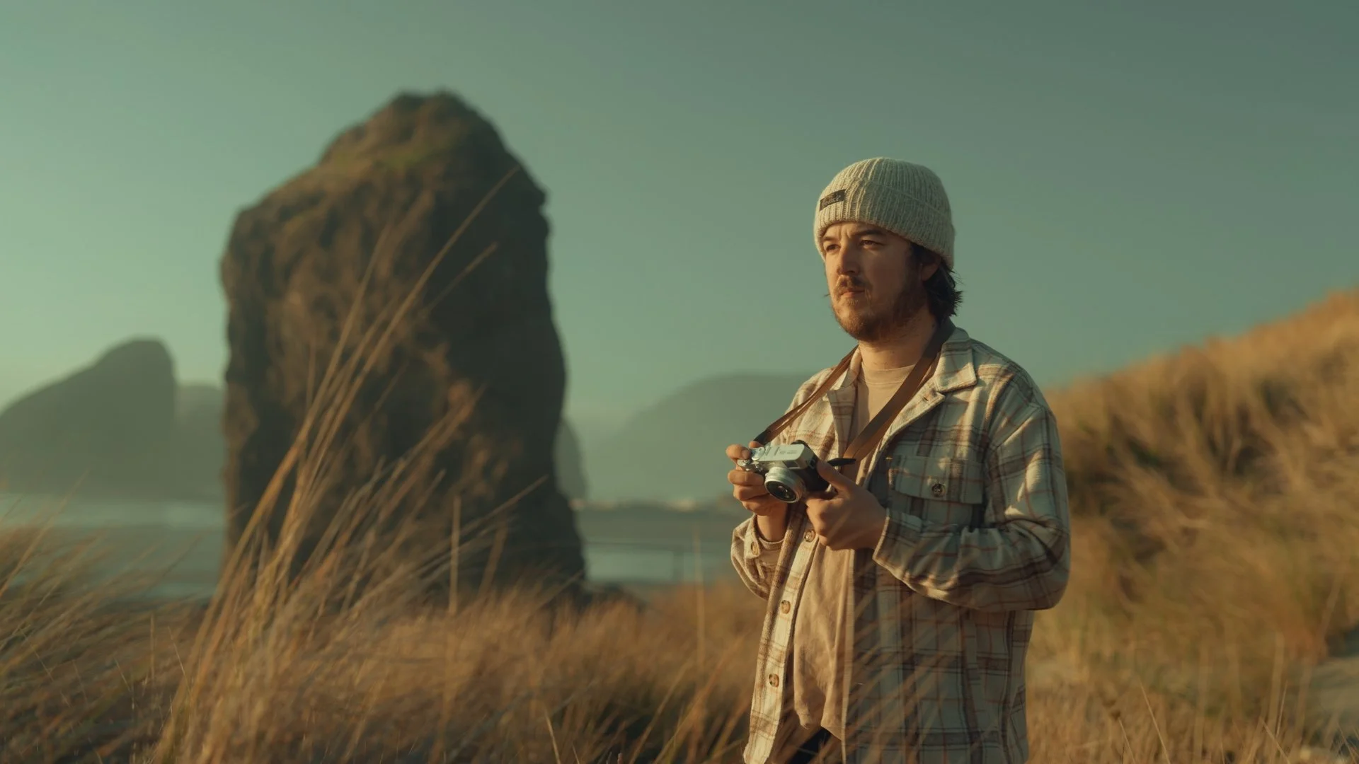 A man with a camera standing on a grassy coastal area, overlooking the ocean with large rock formations in the background, during sunset or sunrise.