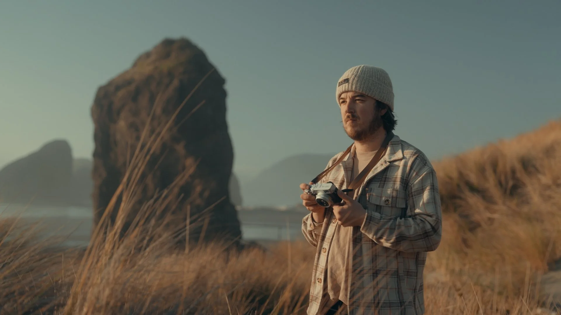 A man in a plaid shirt and knit beanie holding a camera stands outdoors in a grassy area with large rock formations and mountains in the background during daytime.