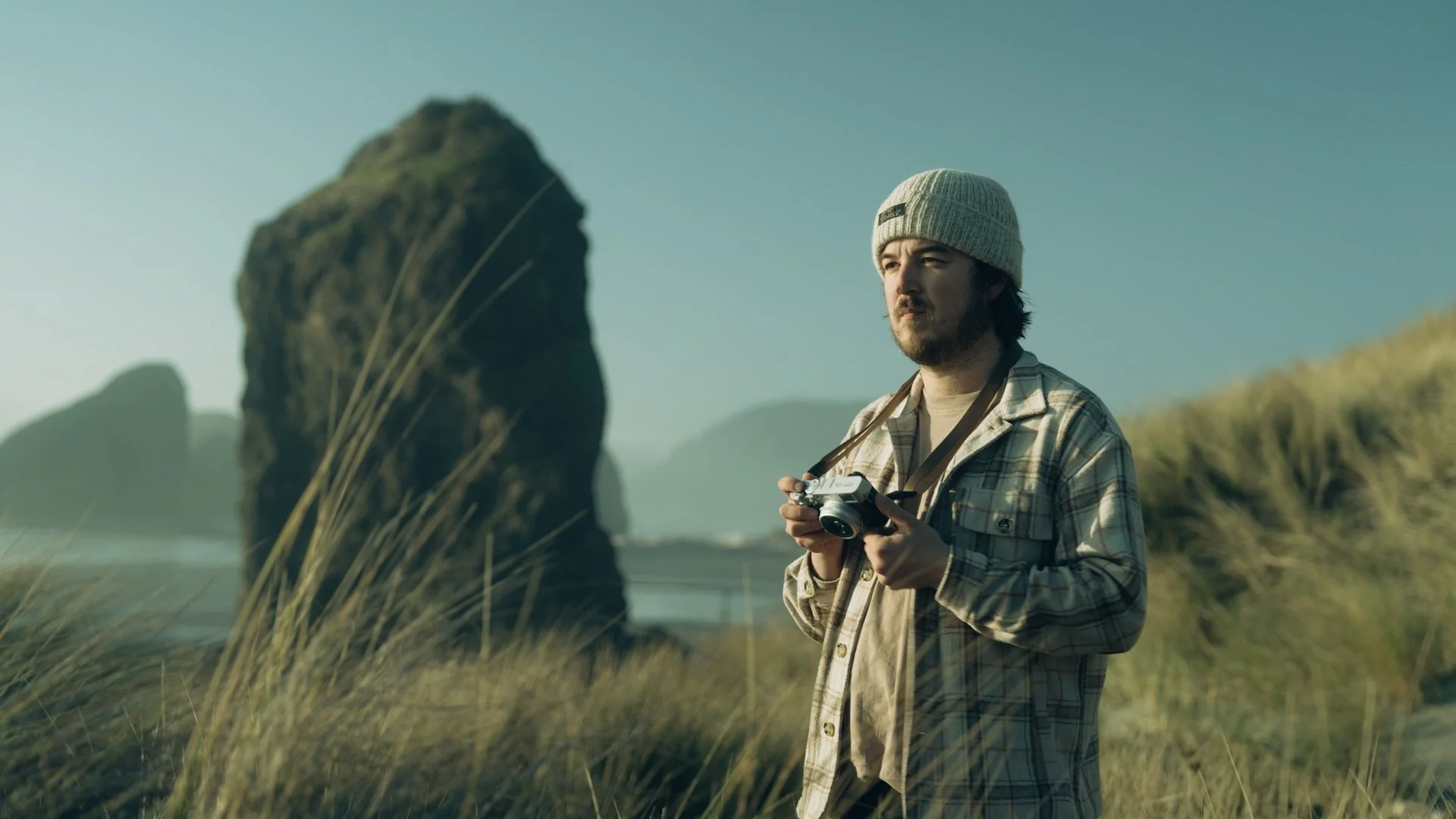 A man holding a camera in a grassy outdoor setting with large rock formations in the background.