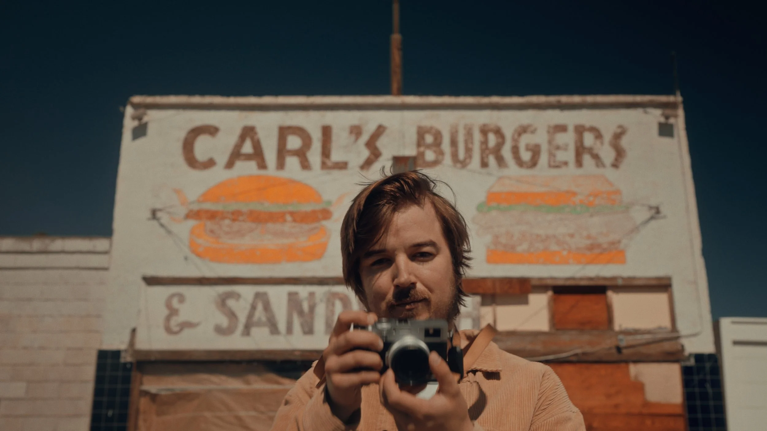 Young man in tan jacket holding camera, standing in front of a painted sign for Carl's Burgers & Sandwiches with illustrations of burgers, at night.