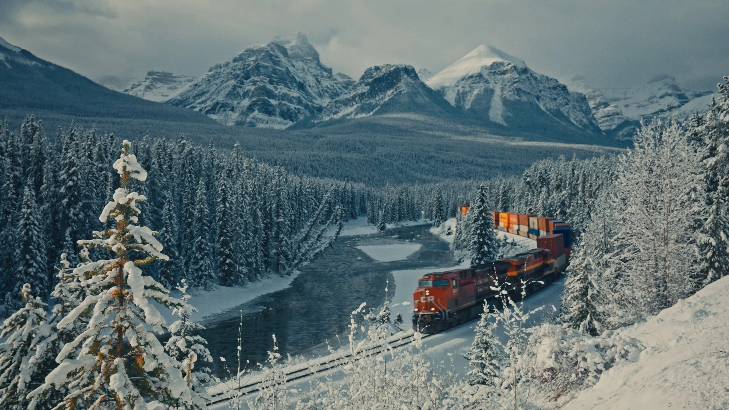 A red freight train with shipping containers is traveling through a snow-covered forested landscape with a river, tall evergreen trees, and snow-capped mountains in the background.