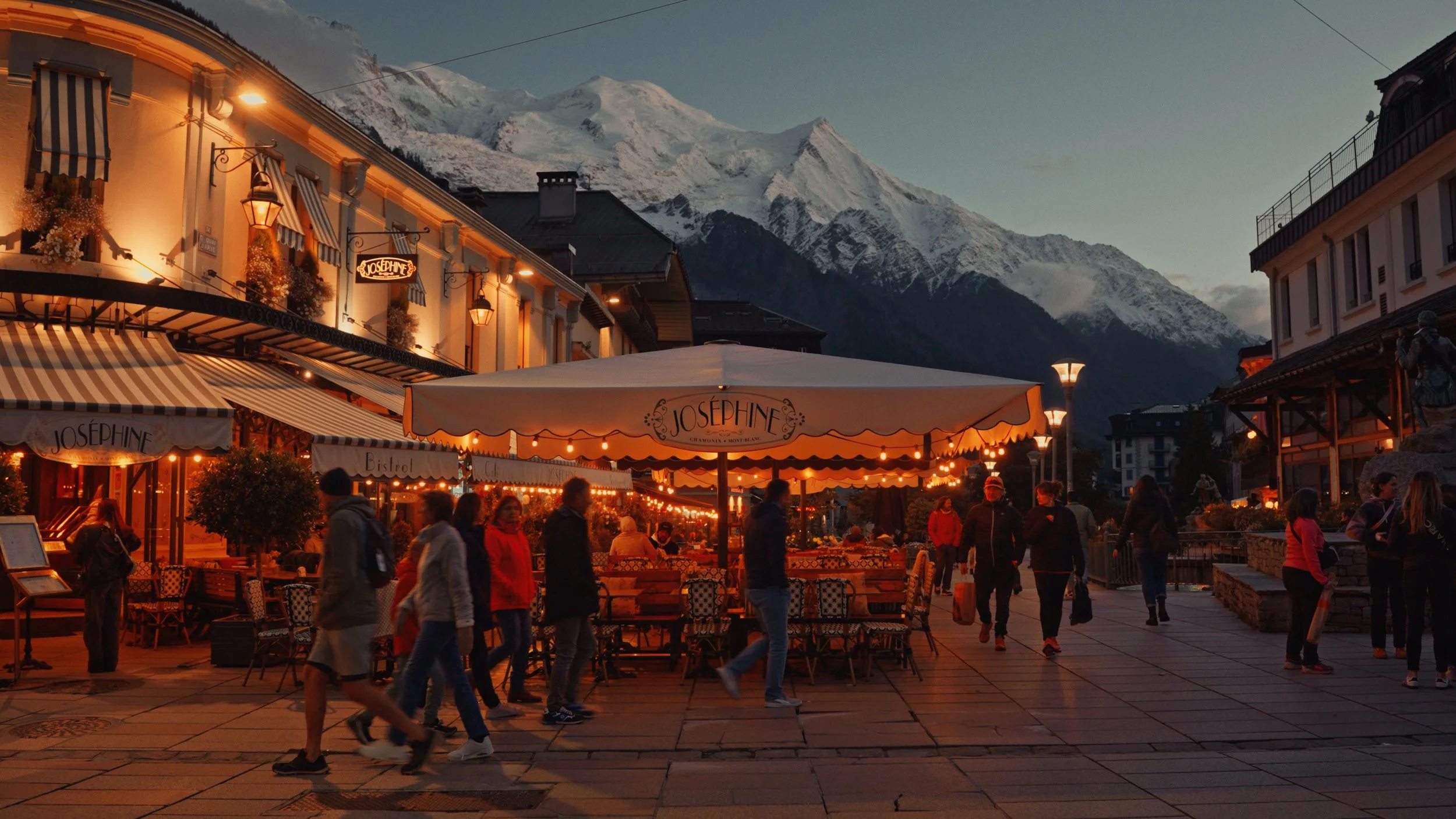People walking along a city street at dusk with illuminated outdoor cafes, mountains in the background, and snow-capped peaks under a darkening sky.