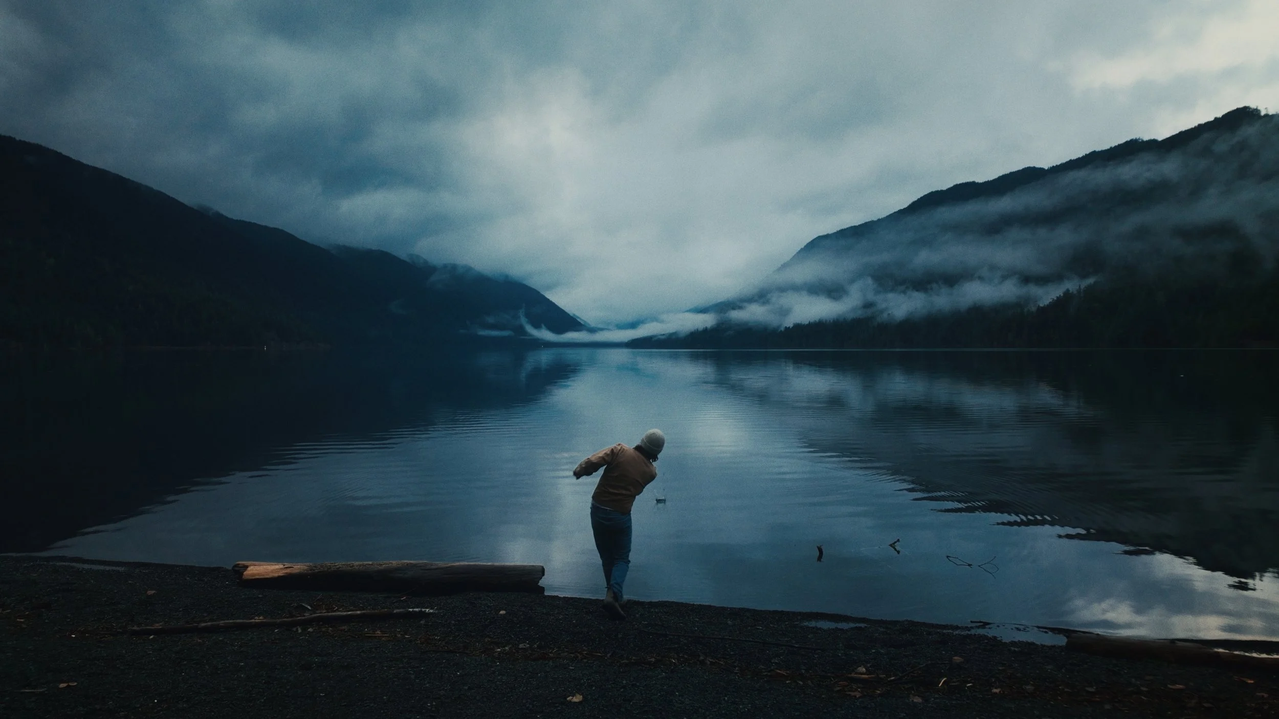 A person standing on a rocky shore by a calm lake with mountains and fog in the background on a cloudy day.