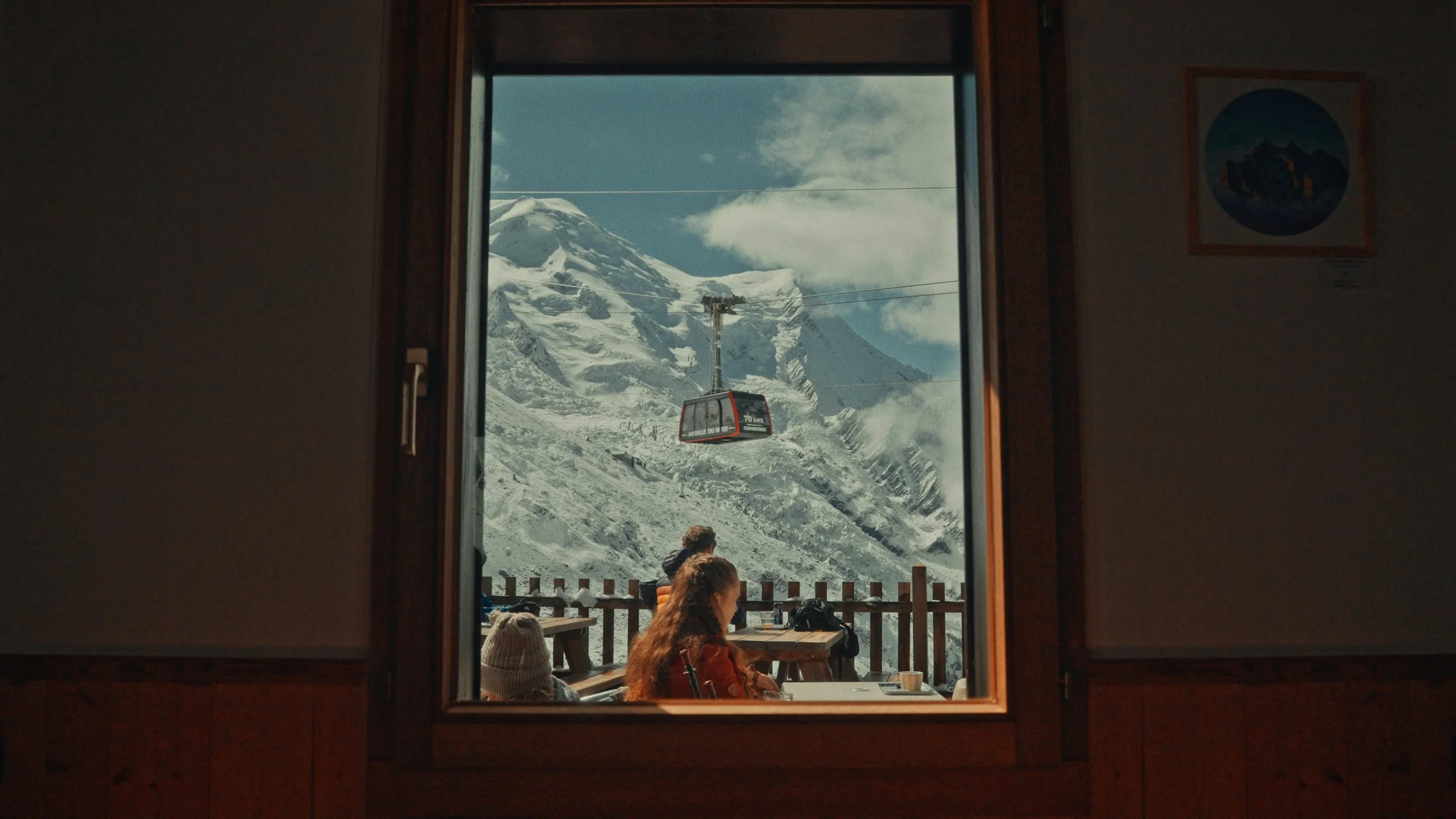 People sitting on a wooden outdoor terrace with snow-capped mountains and a cable car in the background, viewed from inside a building through a window.