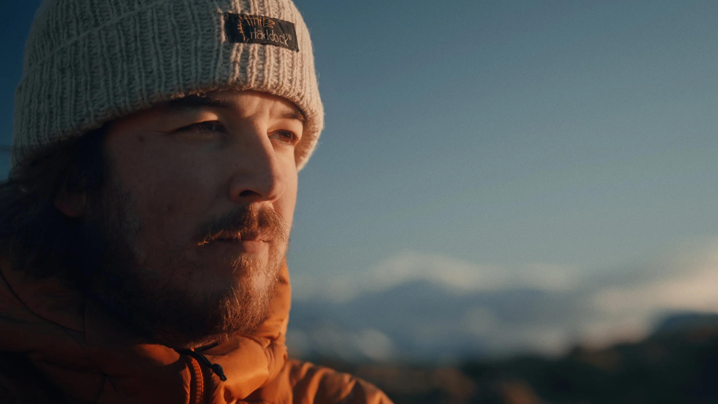 Close-up of a man with a beard wearing a knitted gray beanie and a brown jacket, gazing into the distance during sunset or sunrise.