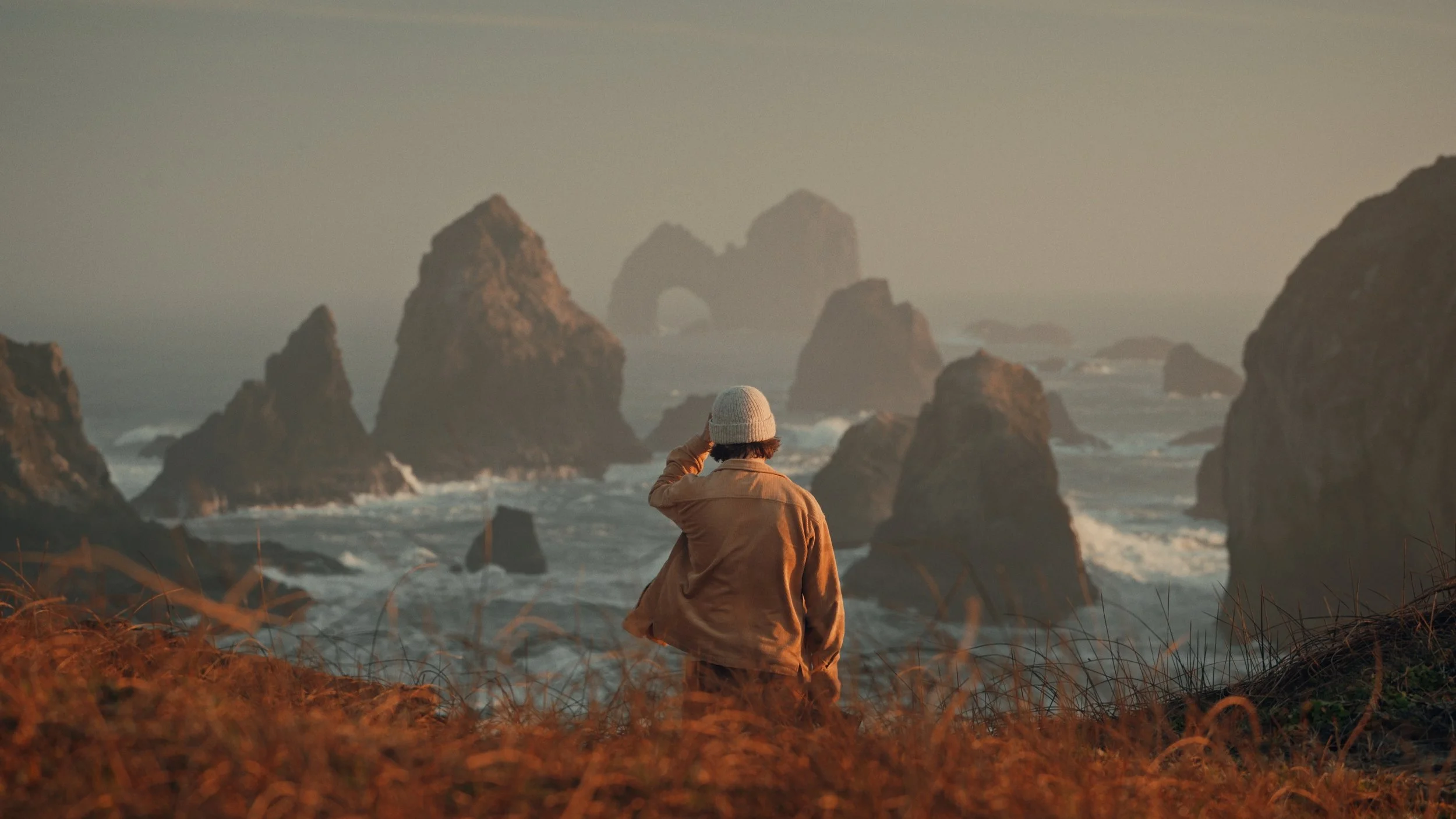 A person wearing a beige jacket and a beanie standing on a grassy hillside facing the ocean with large rocky formations and sea stacks in the distance at sunset.