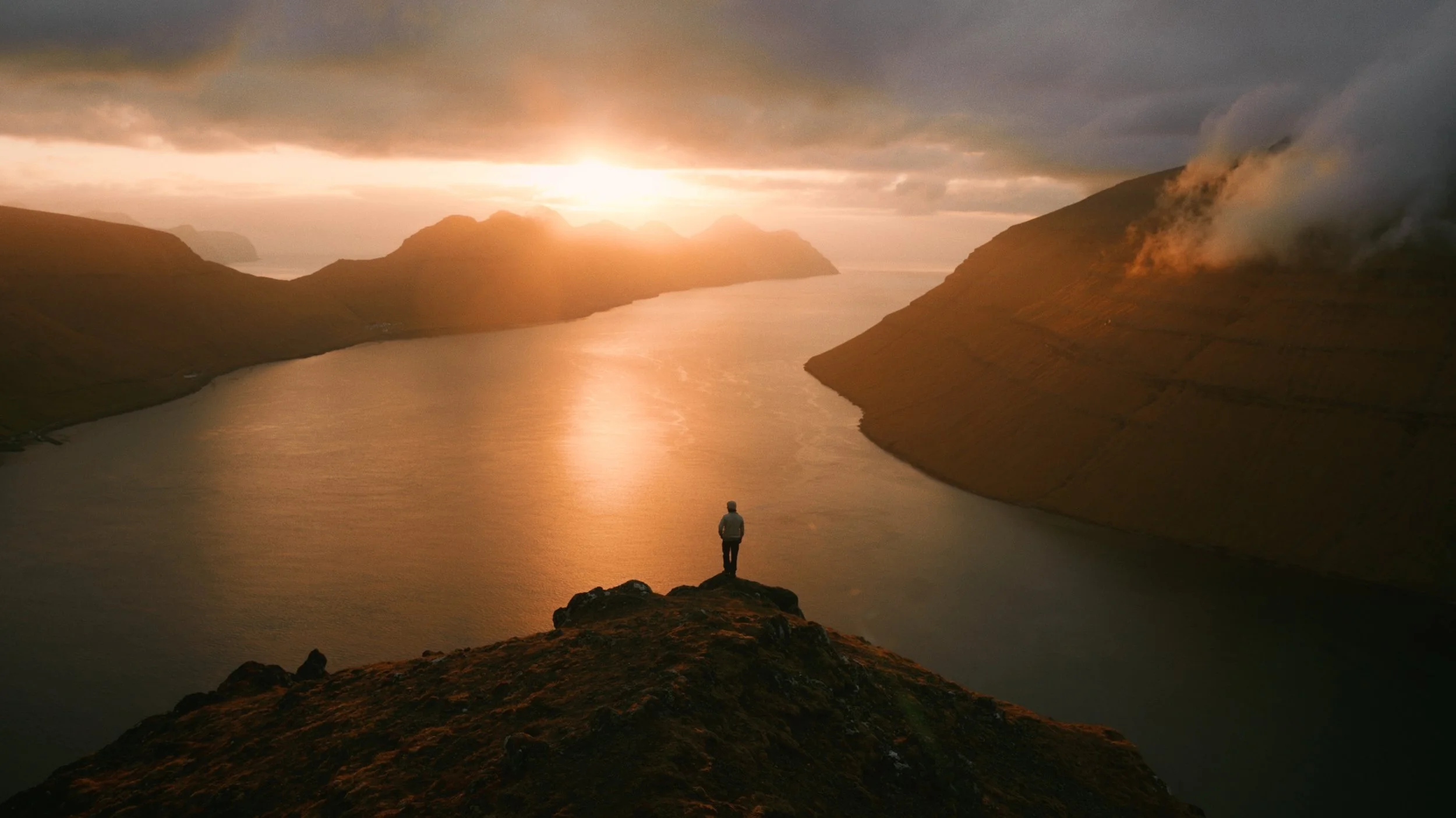 A person standing on a rocky hill overlooking a river at sunset, with mountains and clouds in the distance.