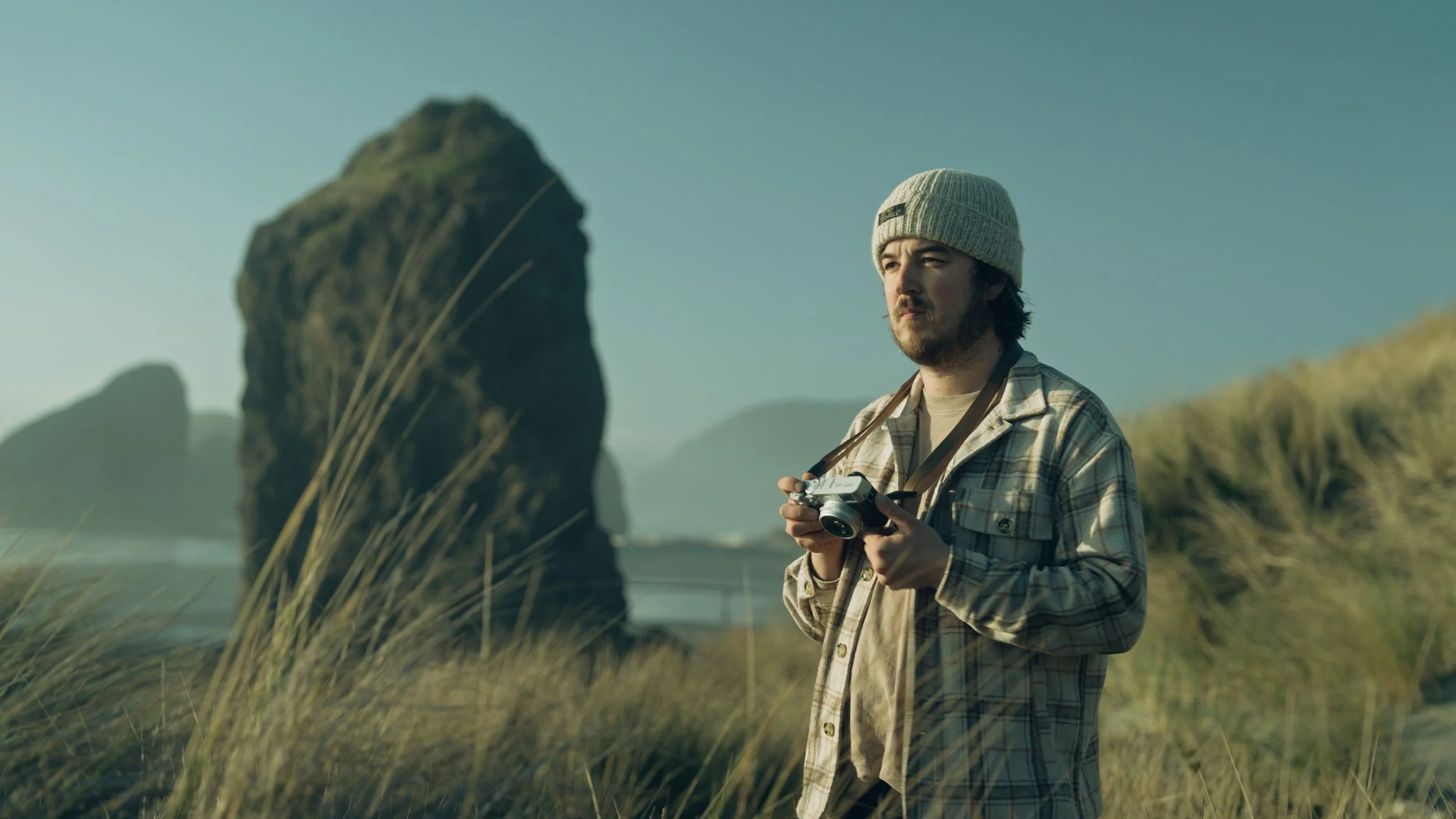 A man with a beard wearing a beanie and plaid shirt holding a vintage camera, standing on a coastal grassy area with large rock formations in the background.
