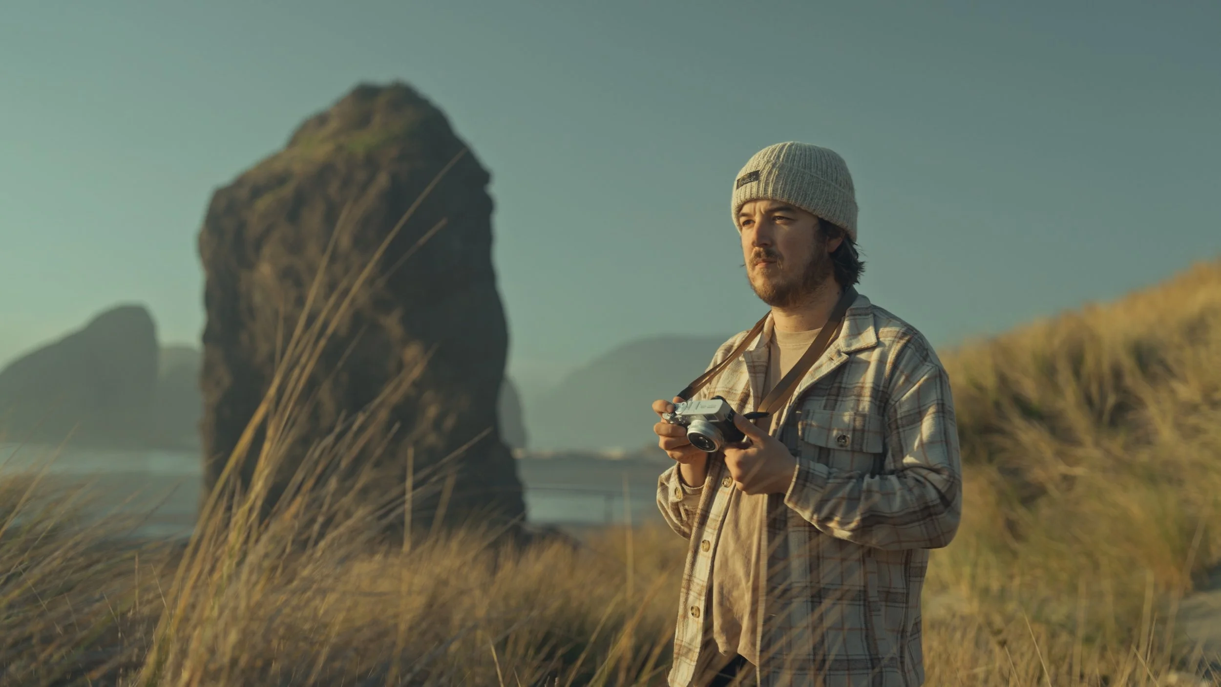 A man with a beard, wearing a gray knit beanie and a beige plaid shirt, stands in a grassy outdoor landscape holding a vintage camera, with large rock formations in the background during sunset.
