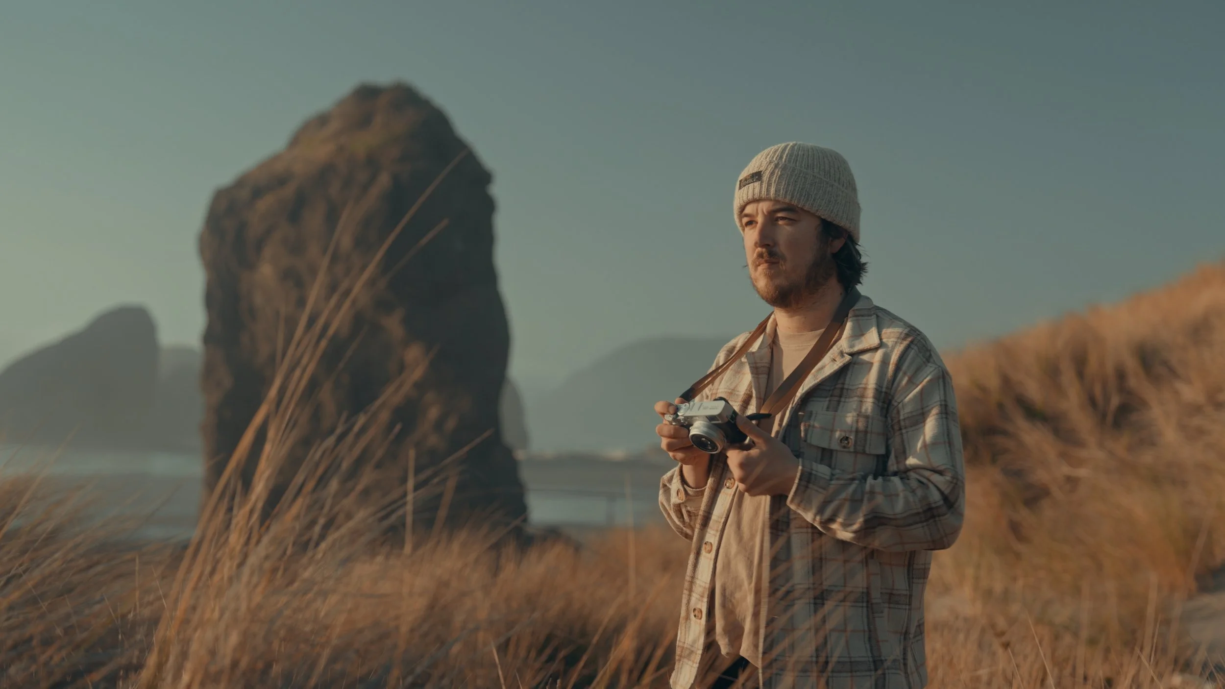 A man standing on a beach with tall grass, holding a camera, wearing a plaid jacket and beanie, with rock formations in the background during sunset.
