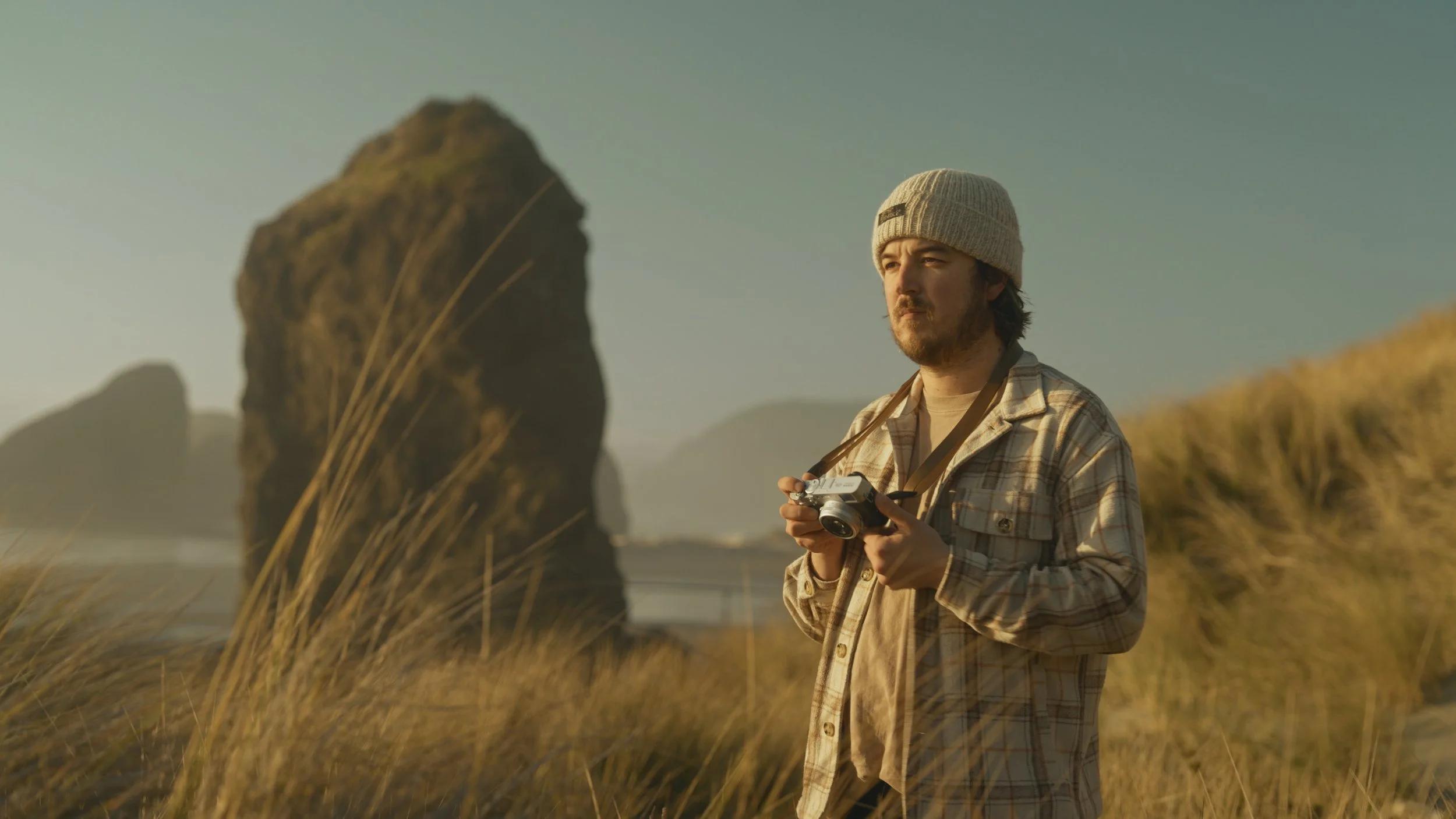 A man stands outdoors in a grassy area, holding a vintage camera, wearing a beige beanie and a plaid shirt, with large rock formations in the background during golden hour.