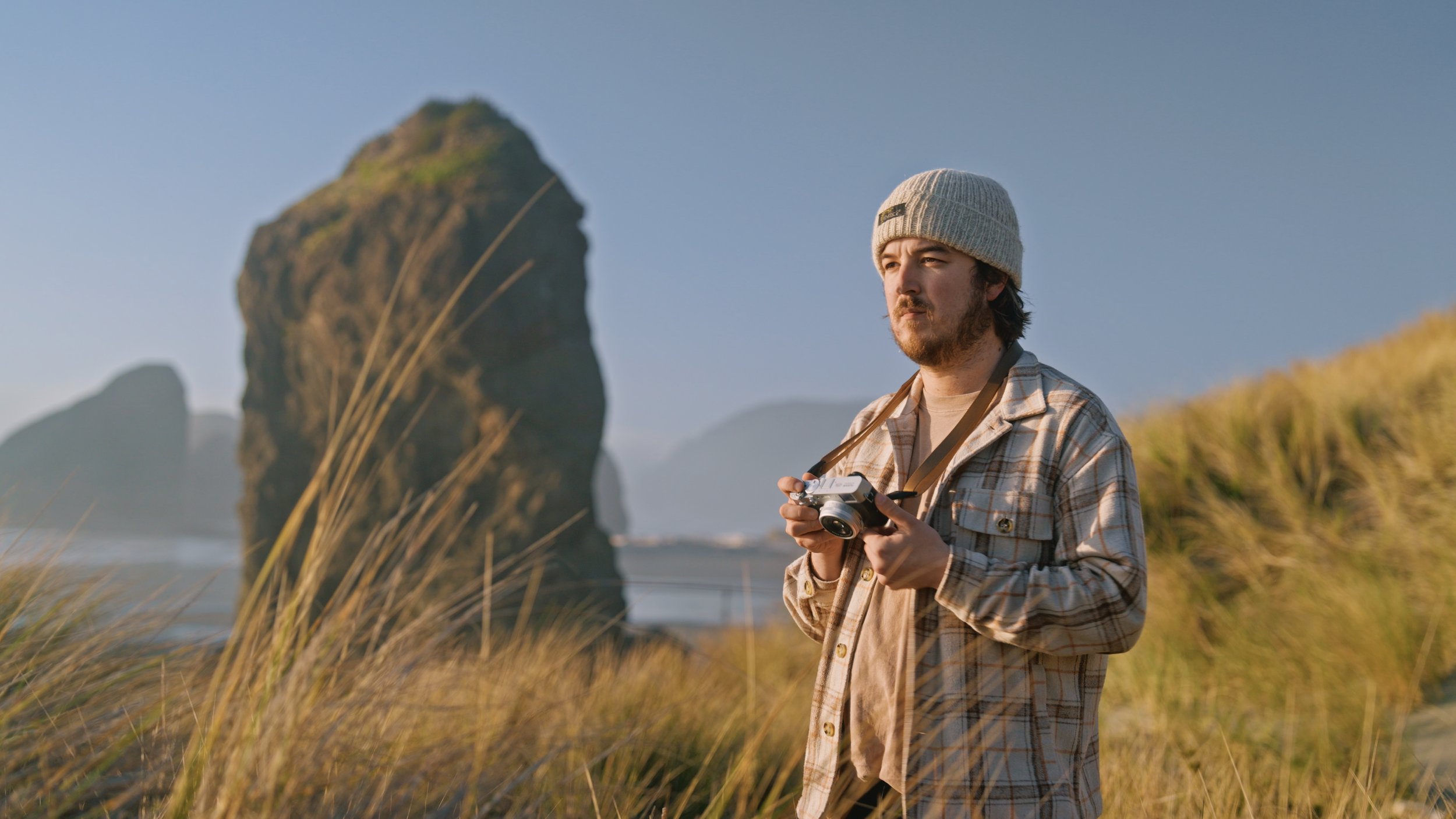 A man standing outdoors on a beach or grassy area, holding a camera and looking into the distance, with large rock formations and dunes in the background during sunset or late afternoon.