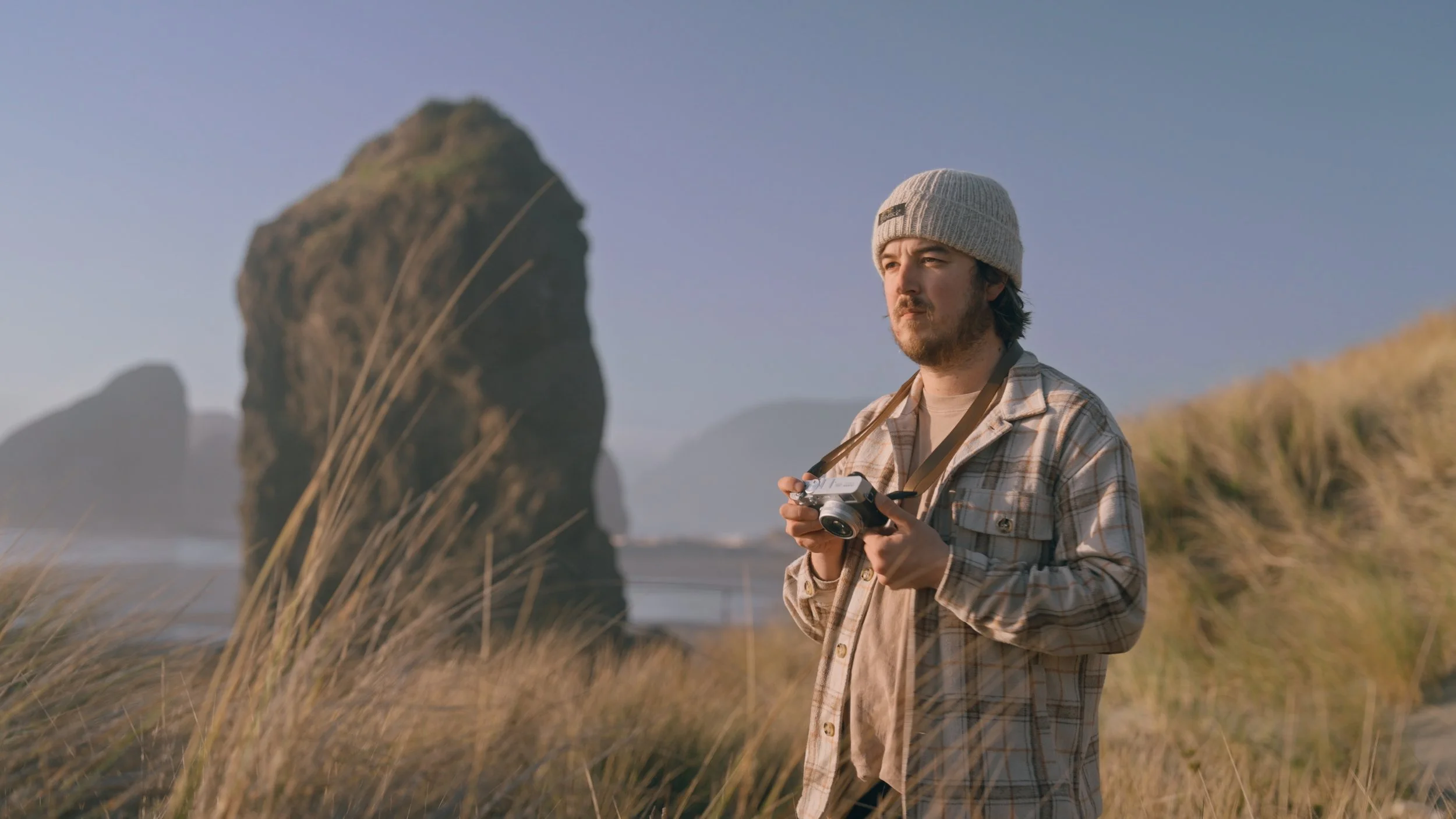 A man wearing a plaid shirt and beige beanie hat holding a camera, standing outdoors on a grassy area near large rocks with a clear sky in the background.