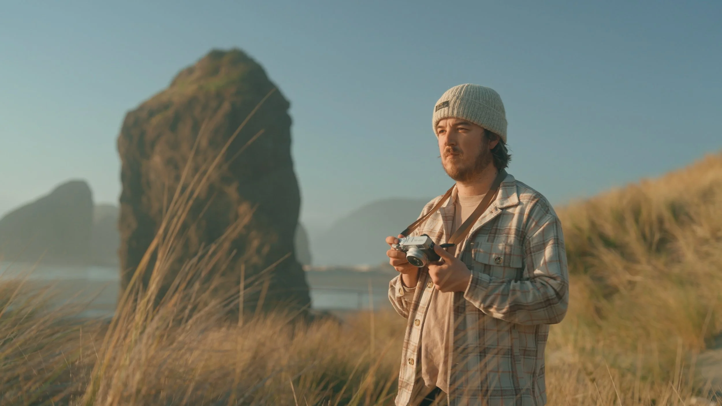 A man with a beard and long hair, wearing a beige beanie and plaid shirt, holding a camera, standing outdoors in a grassy area near large rock formations, during sunset.