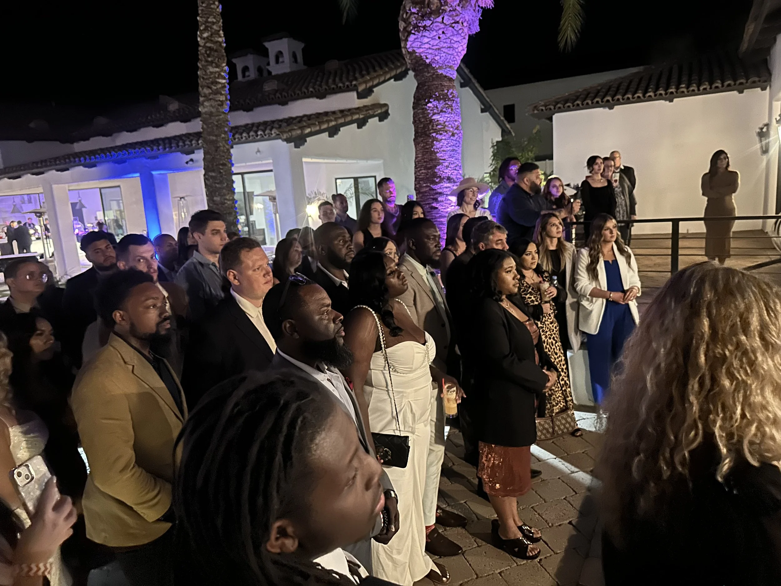 A diverse group of people attending an outdoor event at night, standing and watching attentively. The background features white buildings with tiled roofs, purple lighting on a tree, and some people observing from a balcony.