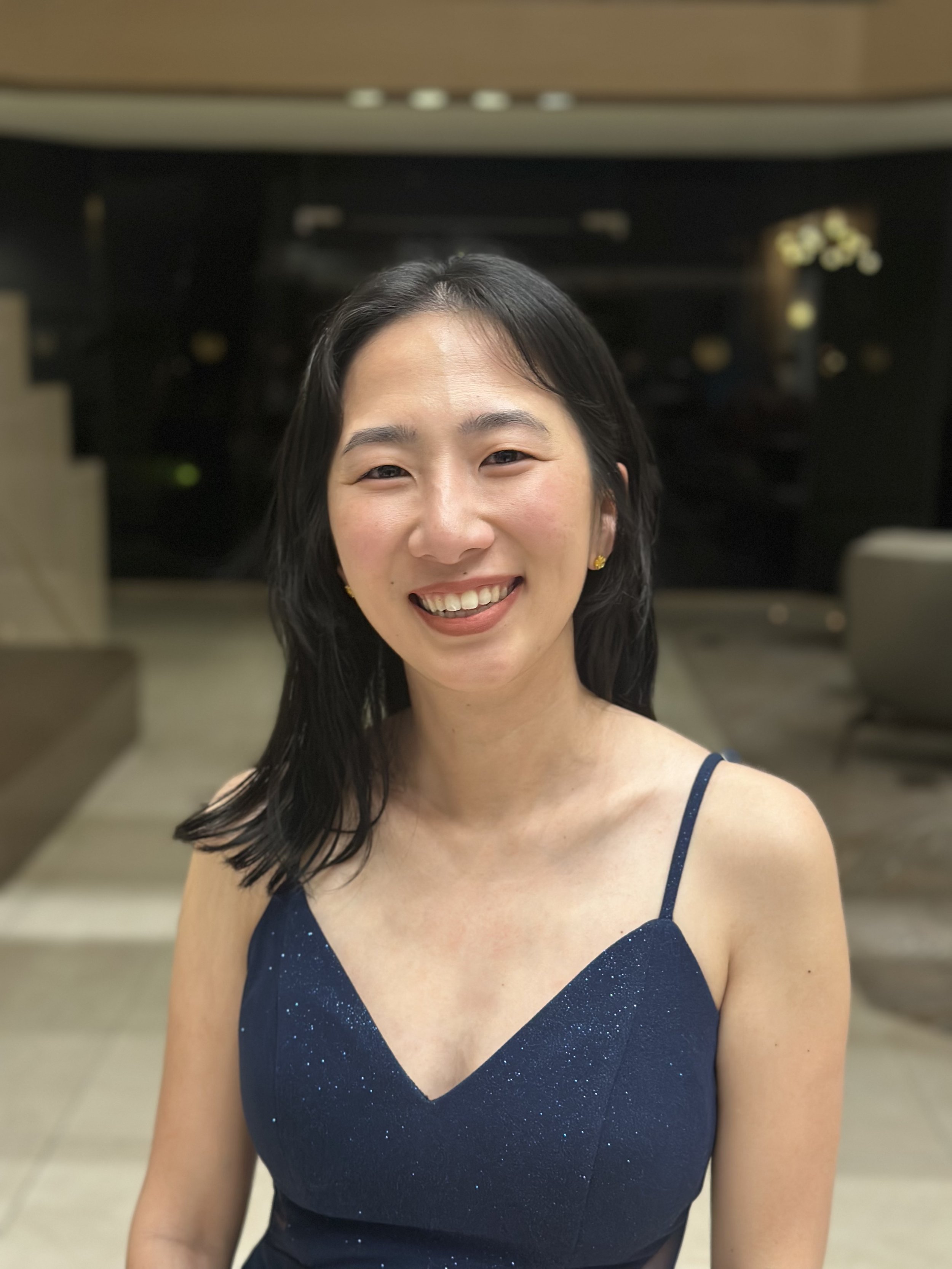 A woman with shoulder-length dark hair, wearing a navy blue dress with thin straps and sparkly details, smiling indoors.