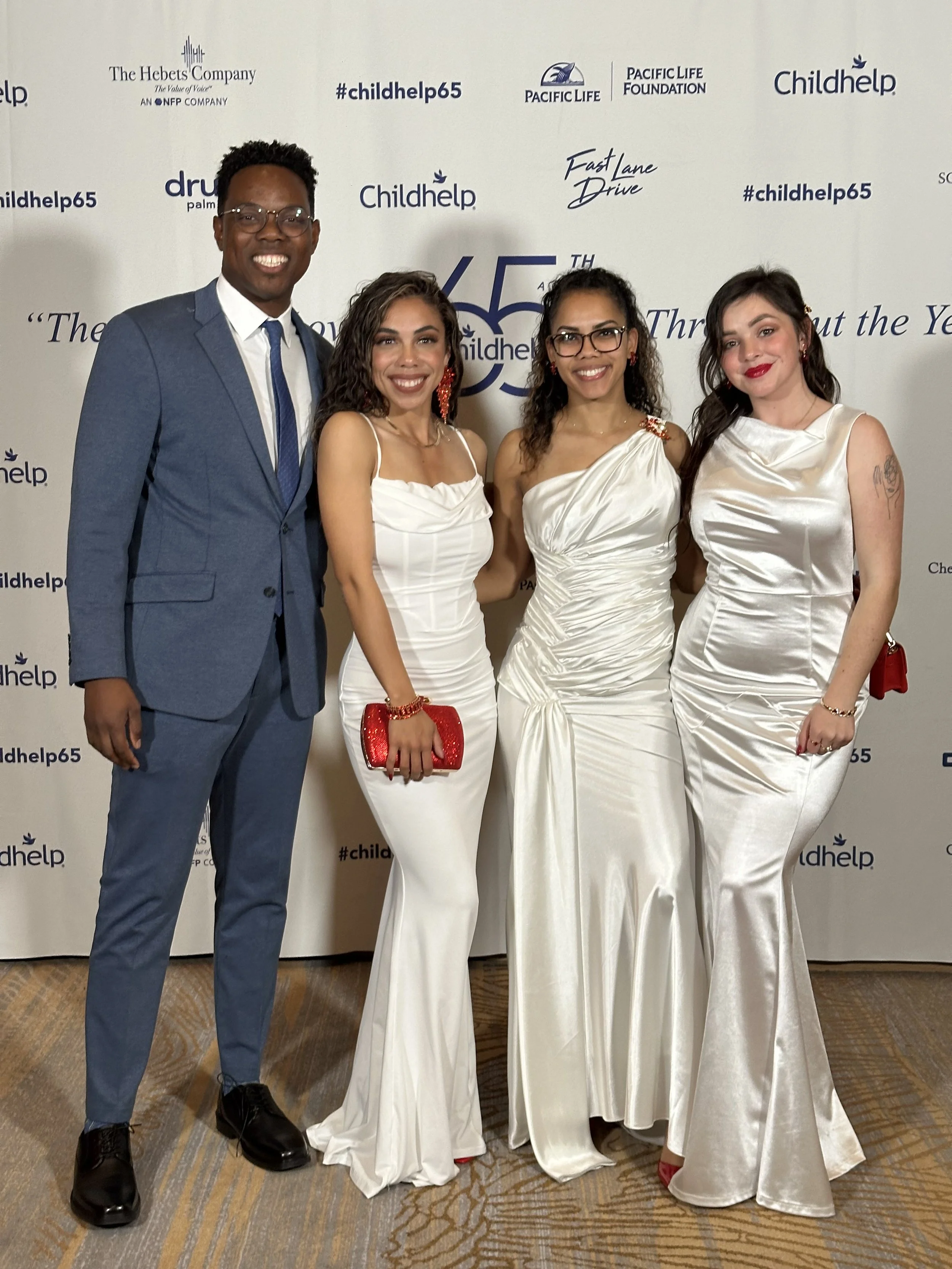 Four individuals dressed in formal attire standing together in front of a backdrop with various logos and hashtags, smiling at the camera.