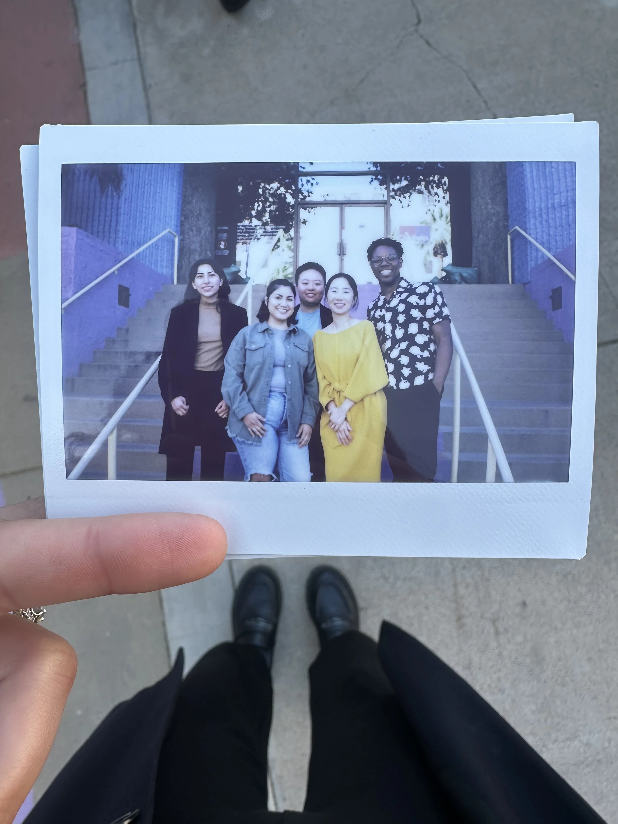 Hand holding a photo of five diverse people standing on stairs outdoors, smiling at the camera, with a building entrance in the background.