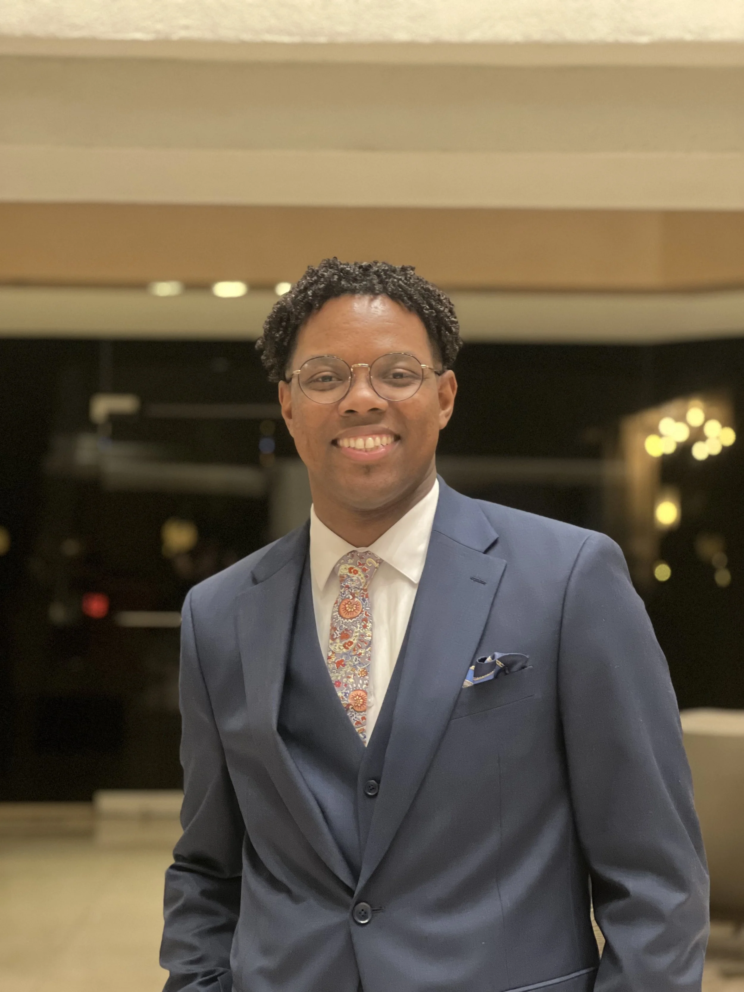 A smiling man in a blue suit, white shirt, and colorful patterned tie, wearing glasses, standing in a warmly lit indoor setting.