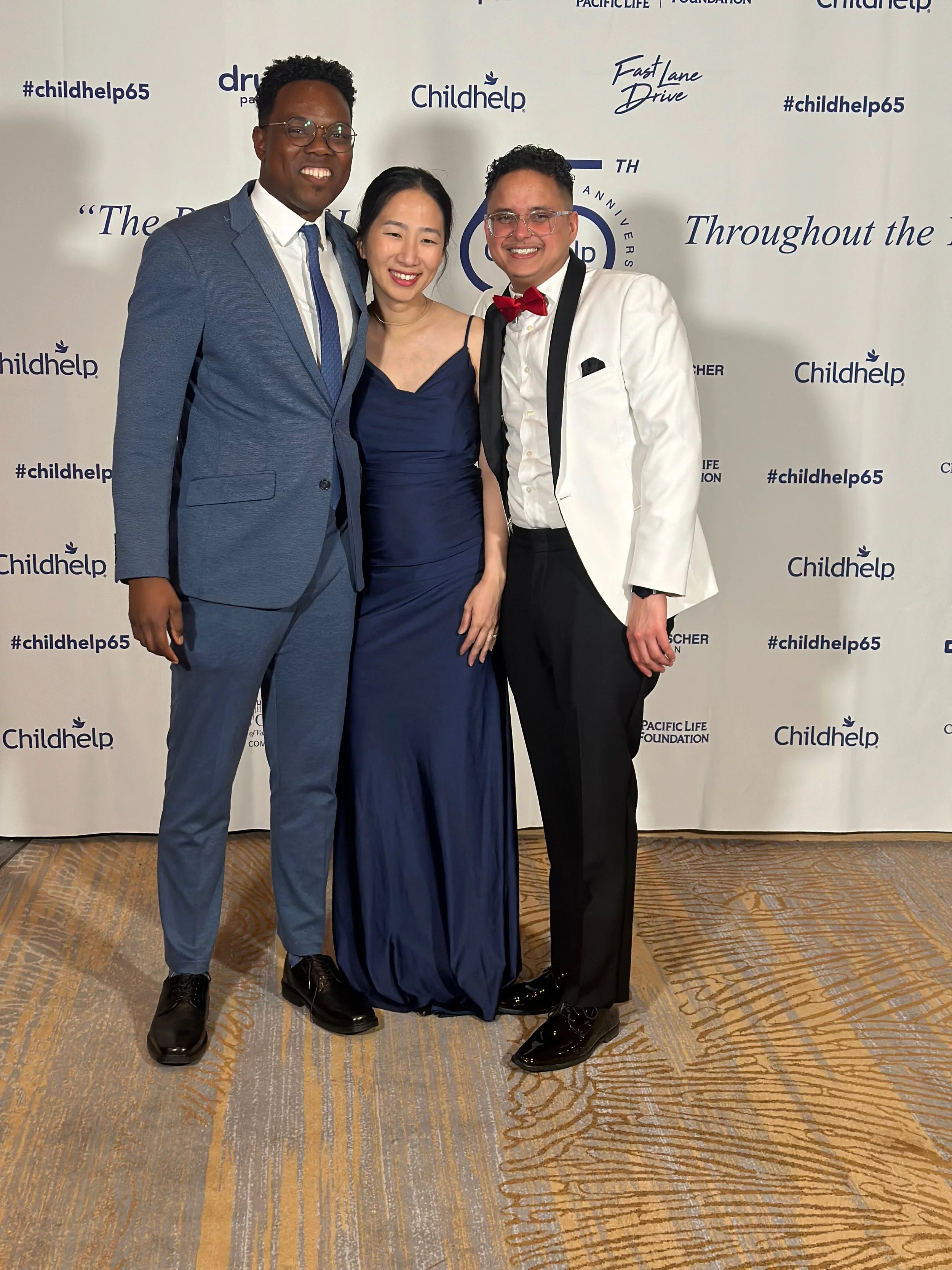 Three people dressed in formal attire standing in front of a white backdrop with logos and text, smiling at the camera.