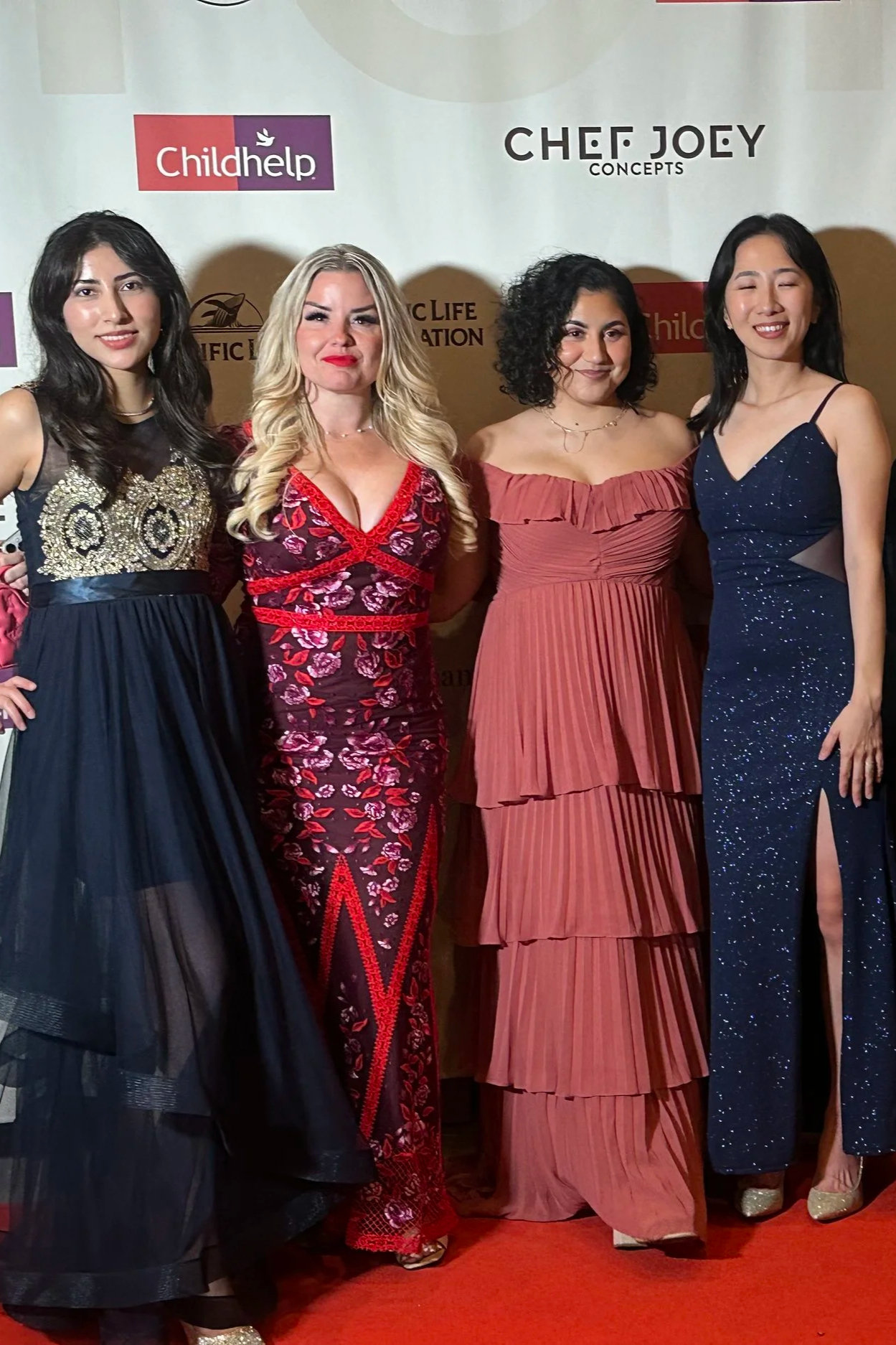 Four women in formal dresses standing together on a red carpet at an event, with a background displaying logos including Childhelp and Chef Joey Concepts.