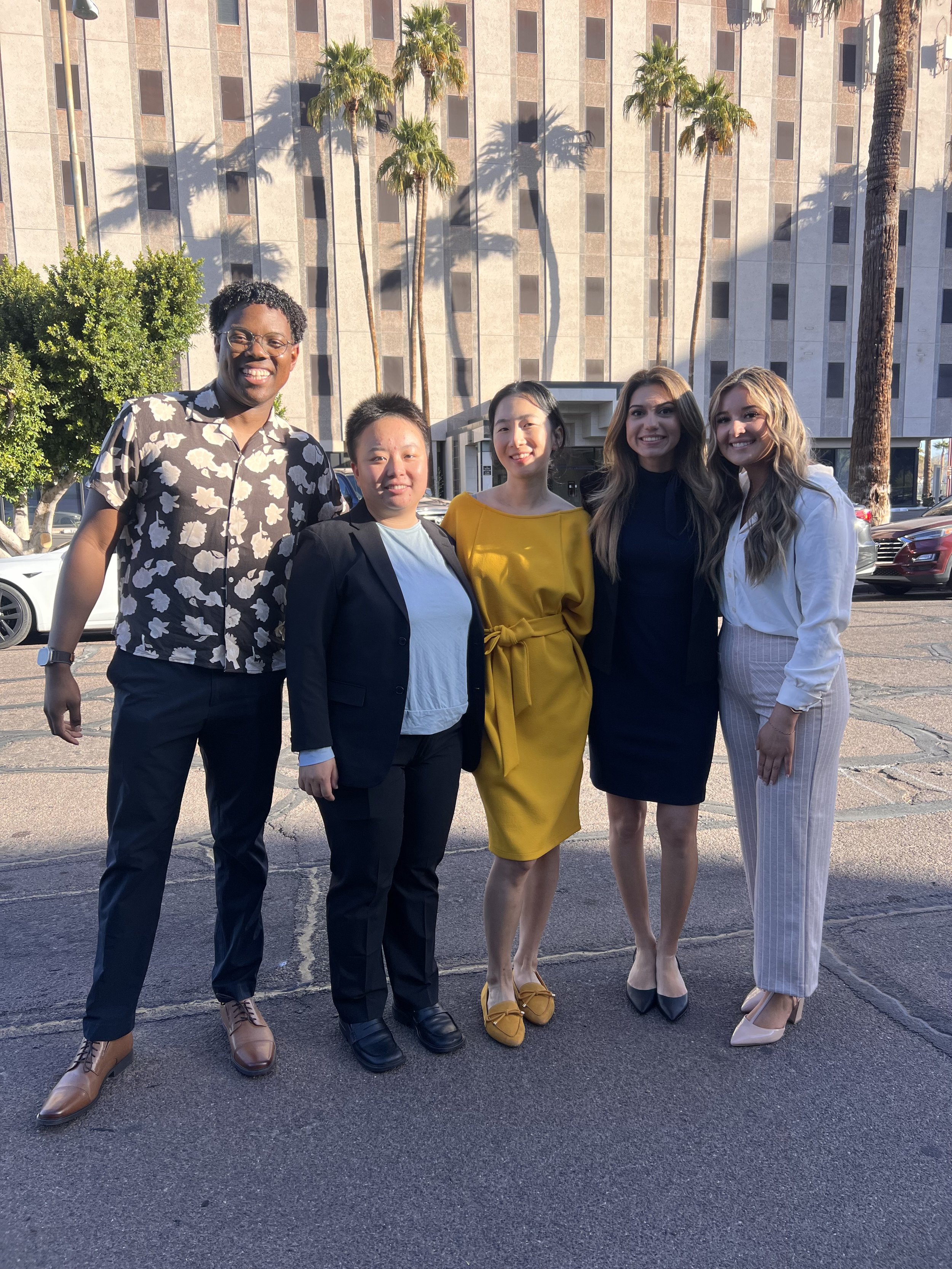 Group of five diverse women standing outdoors in front of palm trees and a building, smiling for the camera.