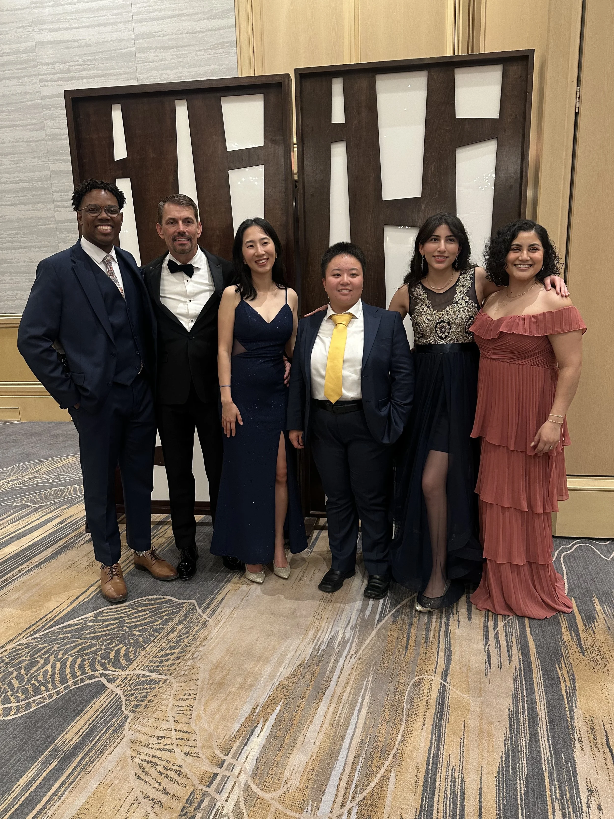 Six people in formal attire standing together in front of large decorative wall art. They are smiling and appear to be at a social event or celebration.