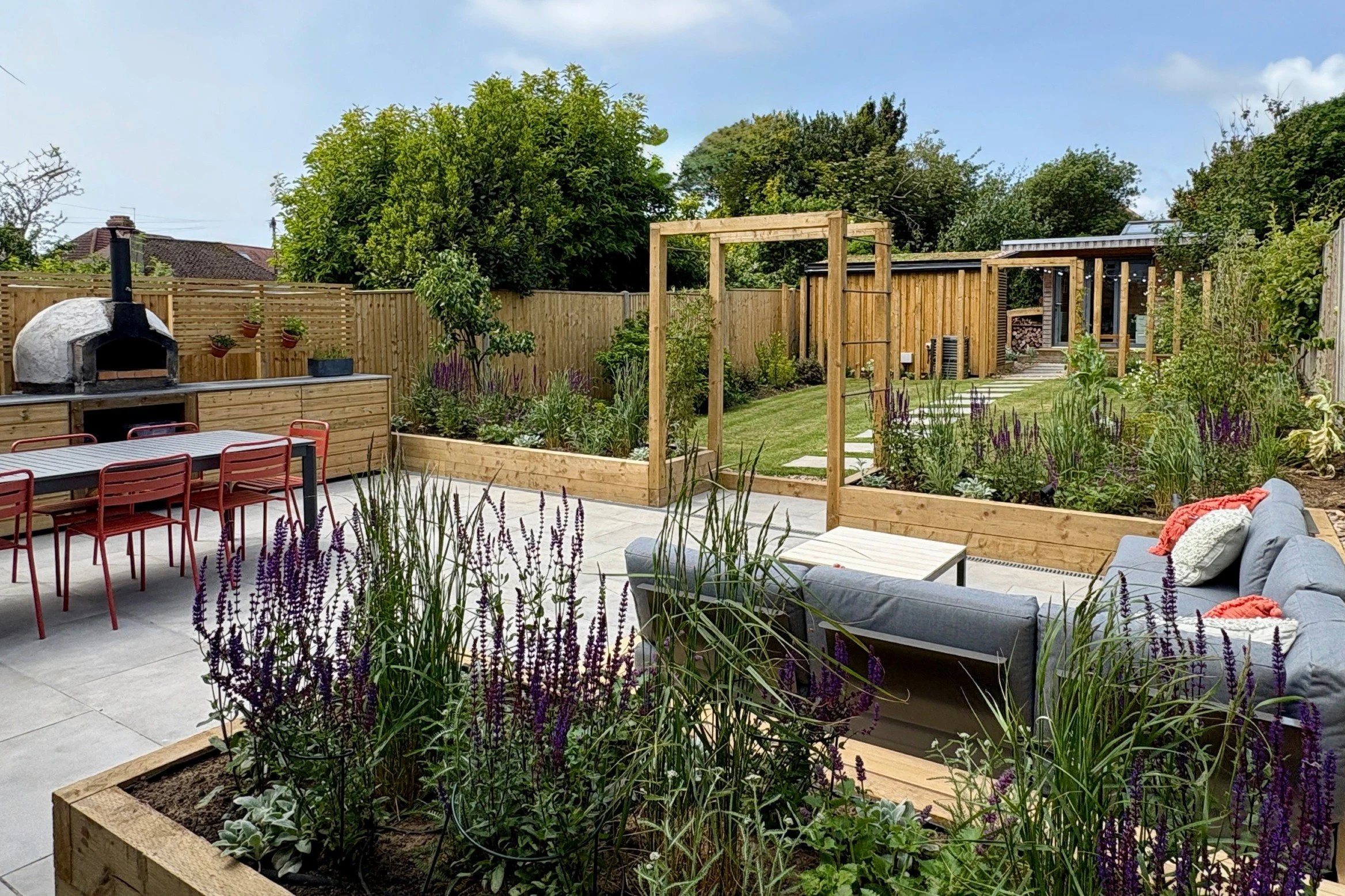 Larger patio in new garden design incorporates outdoor kitchen with pizza oven and barbeque, dining area and sitting area close to the house. Raised beds with perennials and grasses create airy screening and a sense of privacy