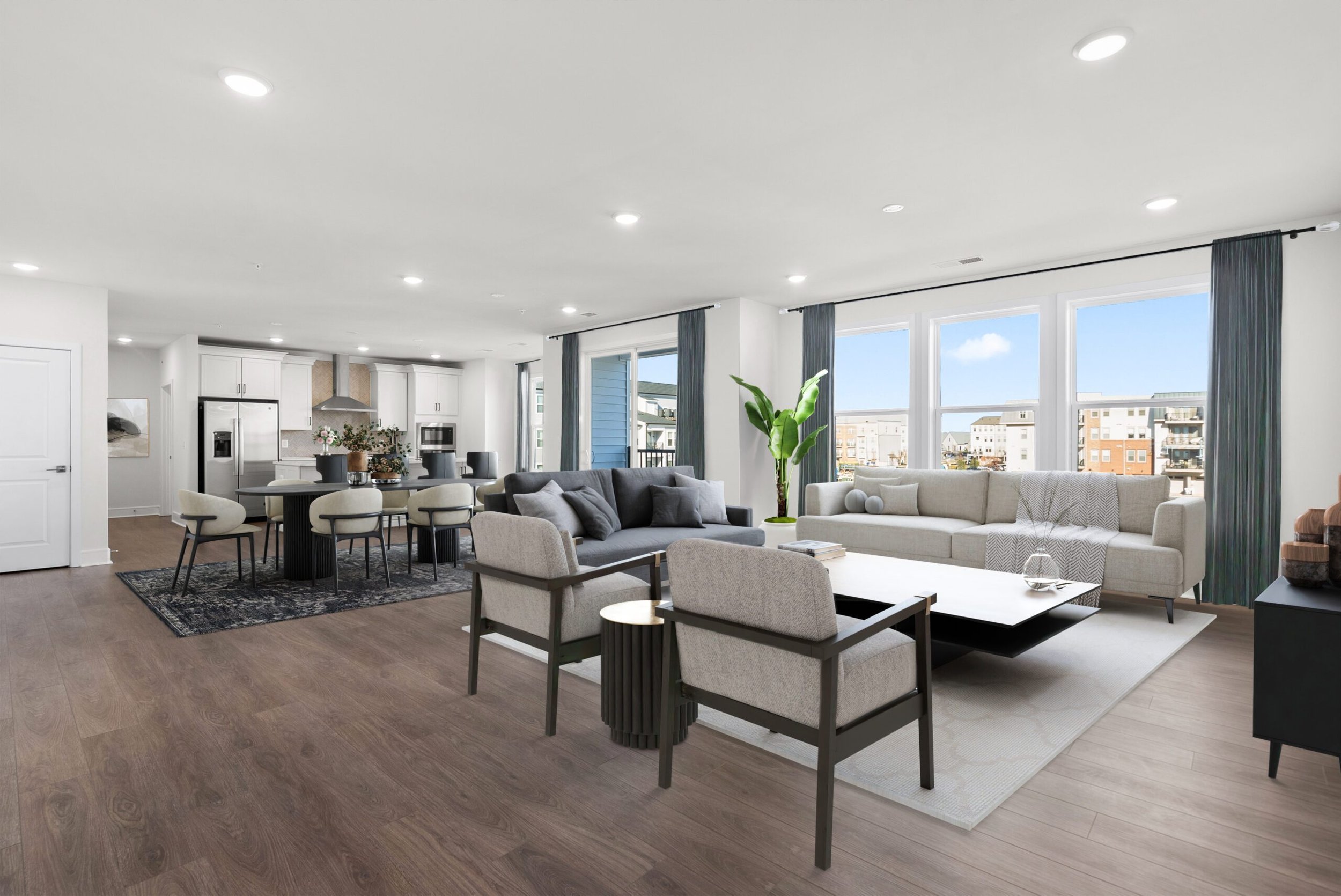 Open-concept living and dining area with large windows, white and gray furniture, plants, wooden flooring, and a view of neighboring buildings.