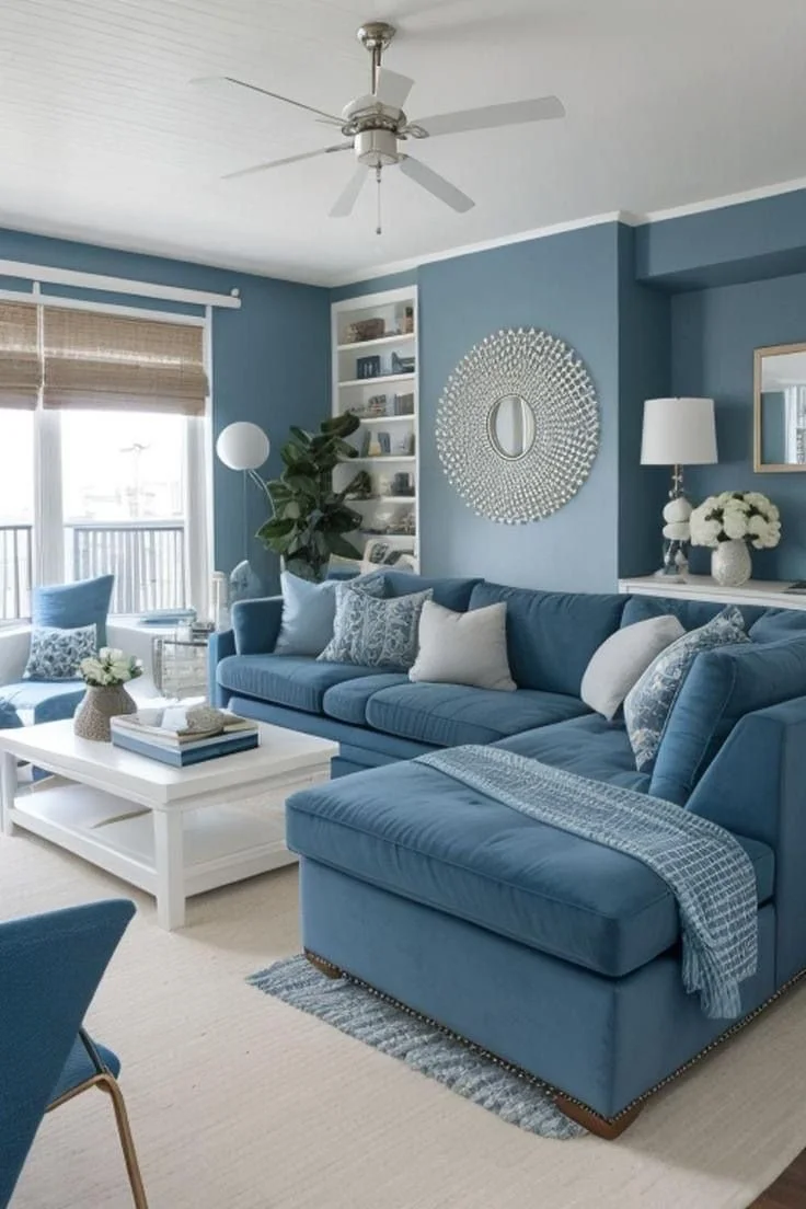 Living room with blue walls and matching blue sectional sofa, white coffee table, large window with bamboo shade, and white ceiling fan.