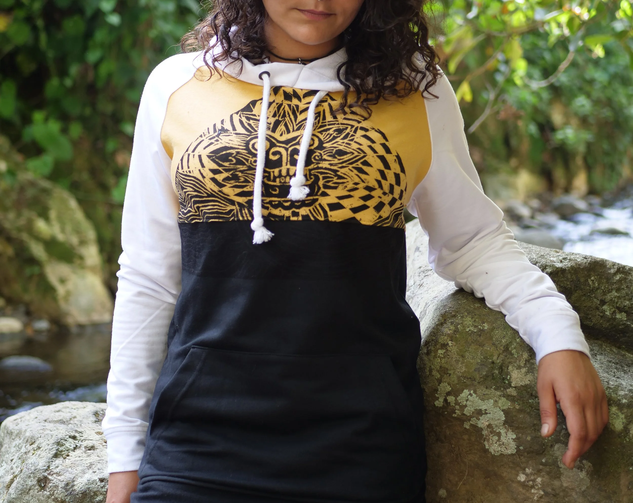 Persona con blusa de manga larga blanca, pantalón negro y con un diseño amarillo y negro en la parte superior, posando junto a una roca en un entorno natural con árboles y agua.