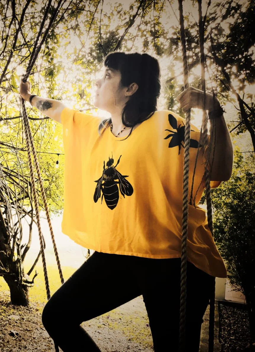 Una mujer con camiseta amarilla con una ilustración de una abeja, sosteniéndose de unas cuerdas en un entorno de naturaleza con árboles y luz solar filtrada entre las hojas.