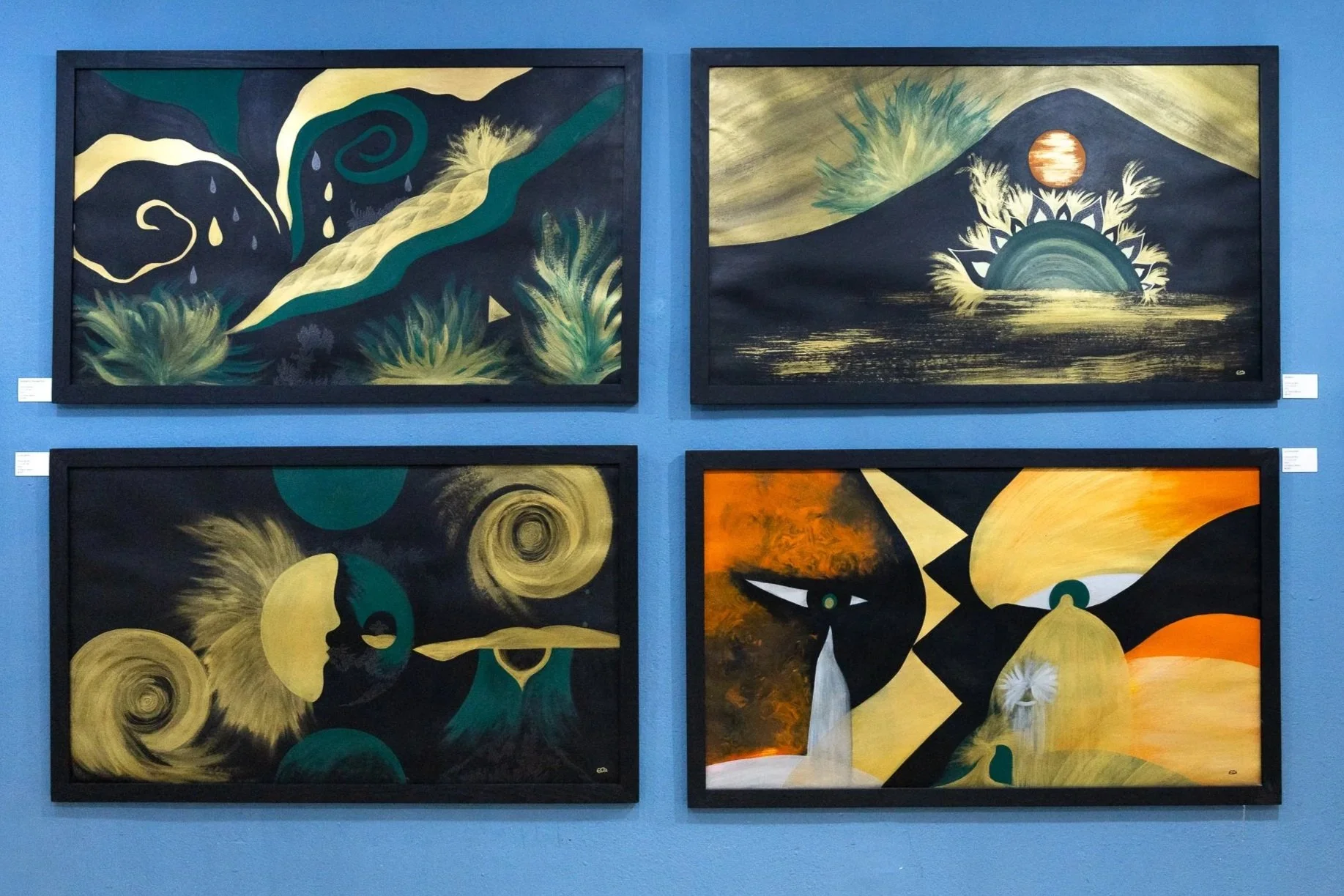Cuatro pinturas abstractas en tonos oscuros con detalles en amarillo, verde y naranja, exhibidas en una pared azul.