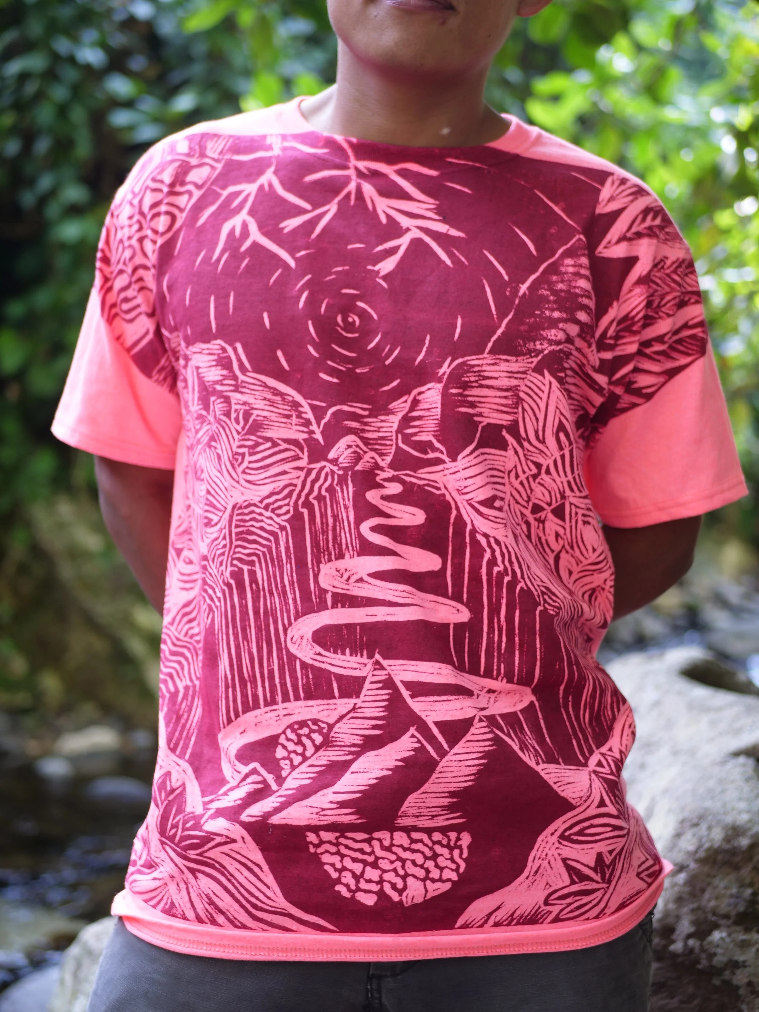 Camisa de color rosa con diseño de montañas, cascadas y un árbol en un entorno natural con árboles verdes de fondo.