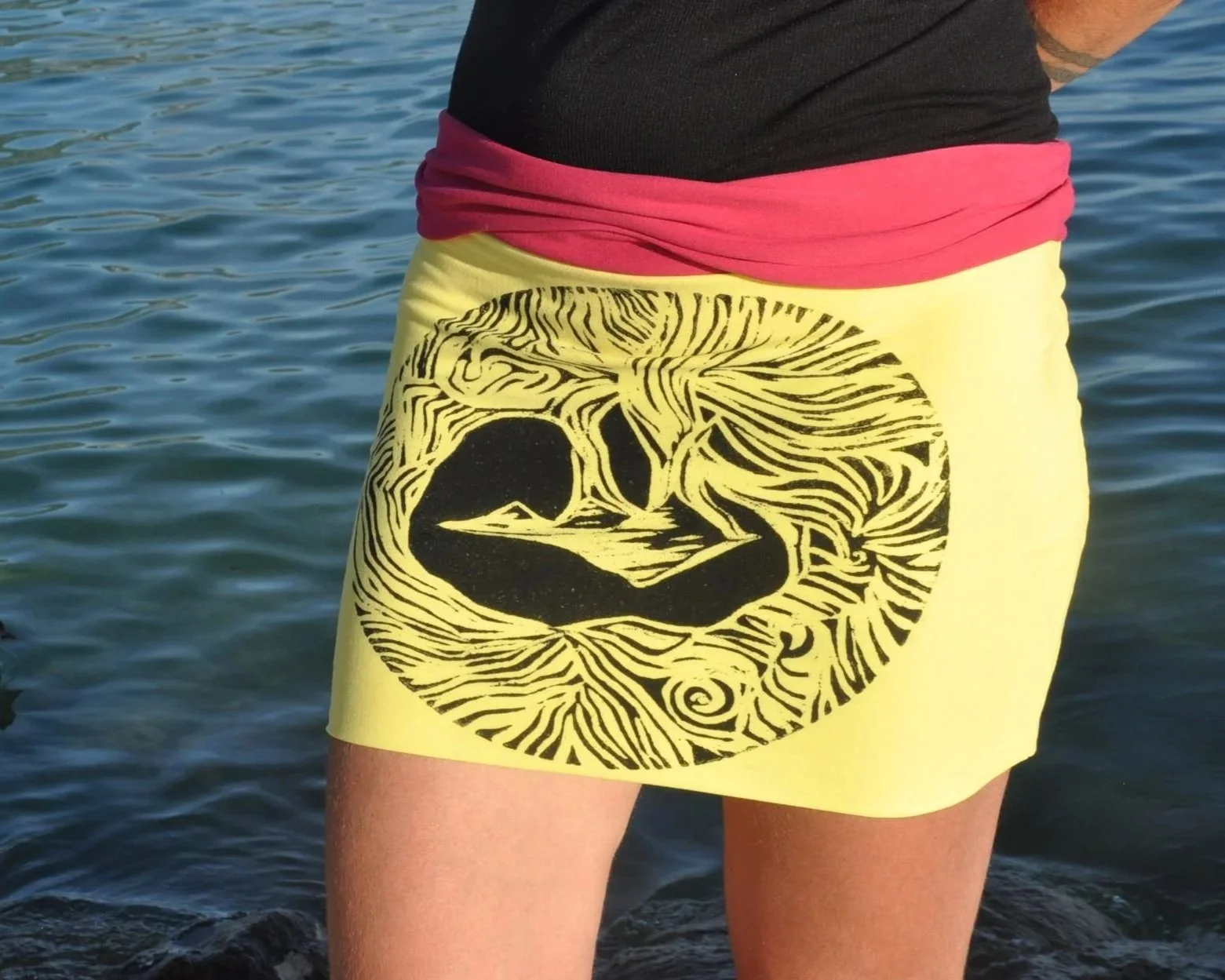 Persona usando falda amarilla con diseño de pato y patrones de líneas negras, frente a agua.