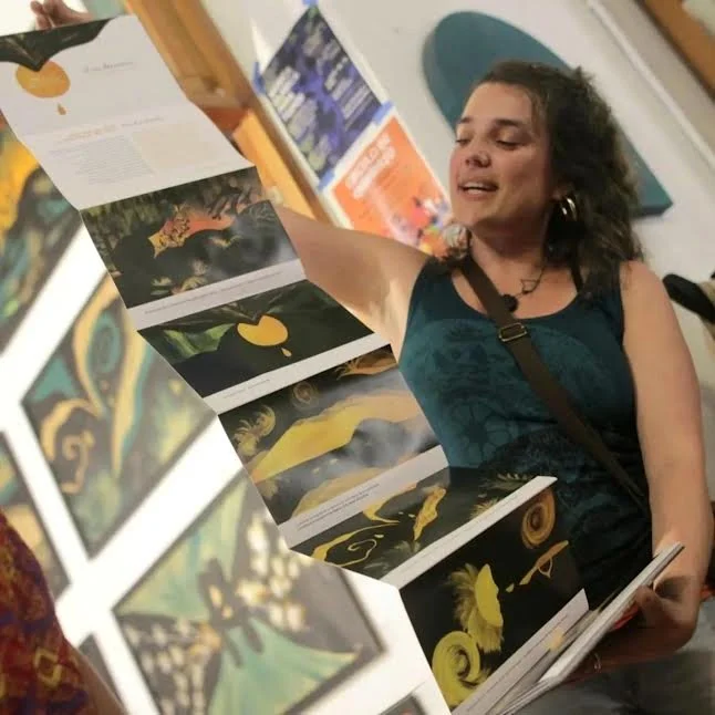 Mujer sonriendo y mostrando un libro desplegado con ilustraciones de arte y naturaleza en una galería o exhibición.