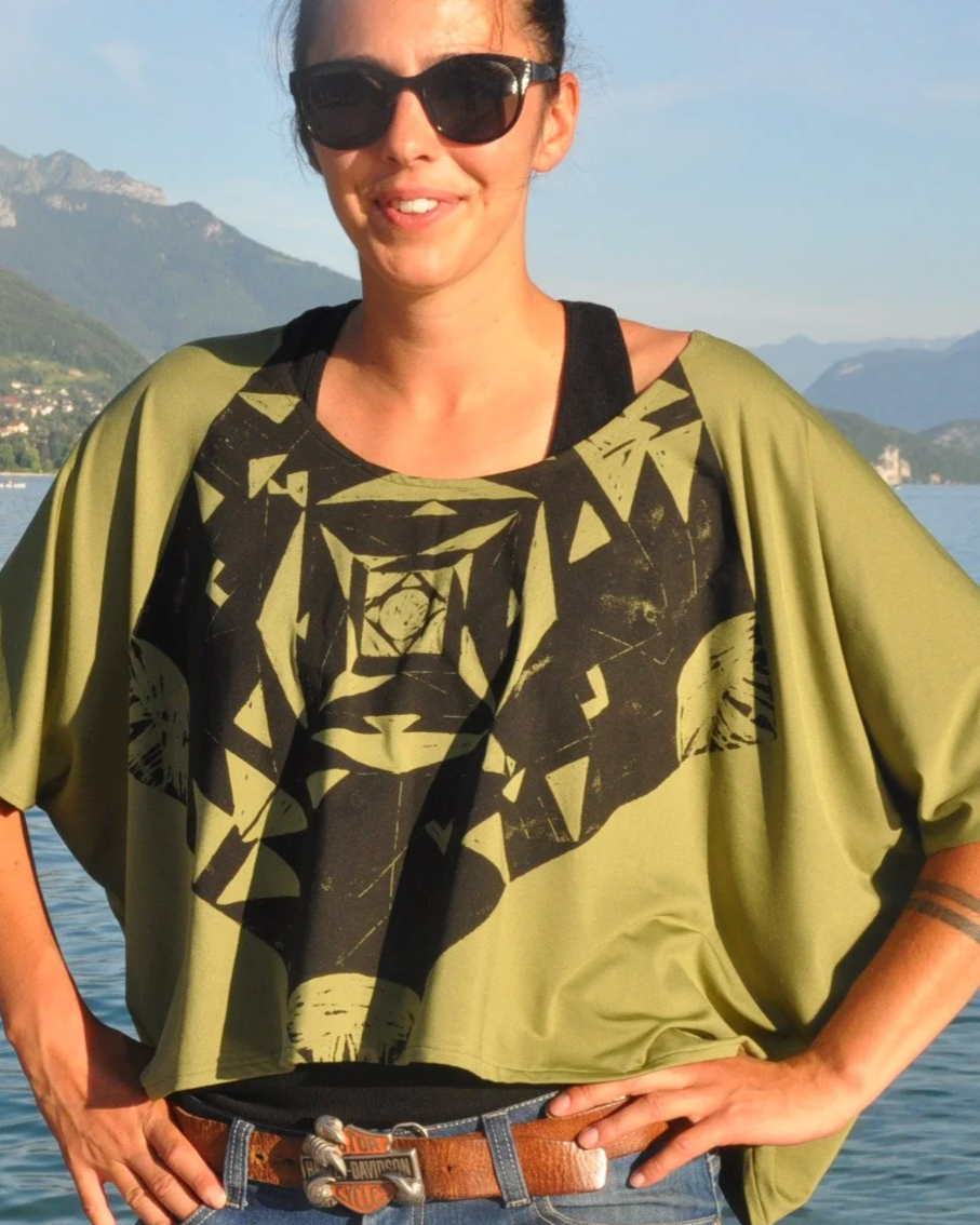 Mujer con gafas de sol en un paisaje montañoso con agua, vestida con camiseta con estampado y chaqueta verde.