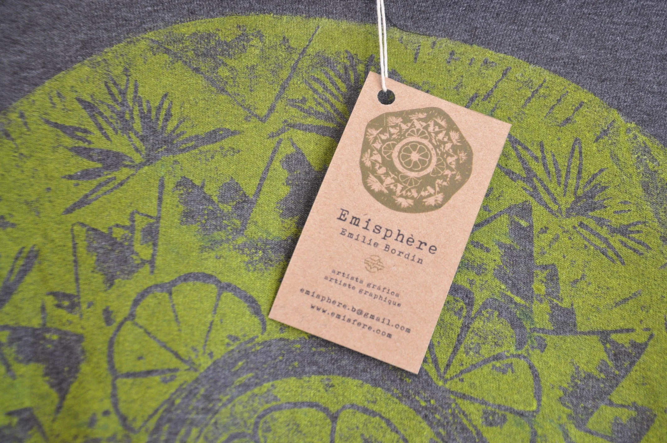 Etiqueta de cartón con detalles gráficos y texto que identifica a Emisphere y a la artista Emile Bordin, colocada sobre tela gris con un diseño en verde y gris.