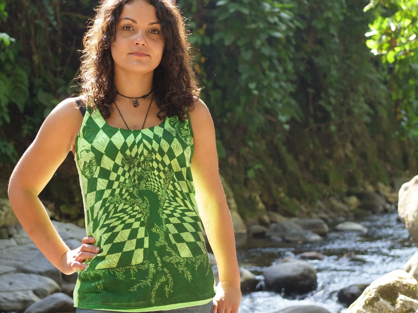 Una mujer con cabello rizado oscuro, vestida con una camiseta verde con un patrón de un laberinto y un árbol en el centro, de pie junto a un río con rocas y vegetación verde en el fondo.