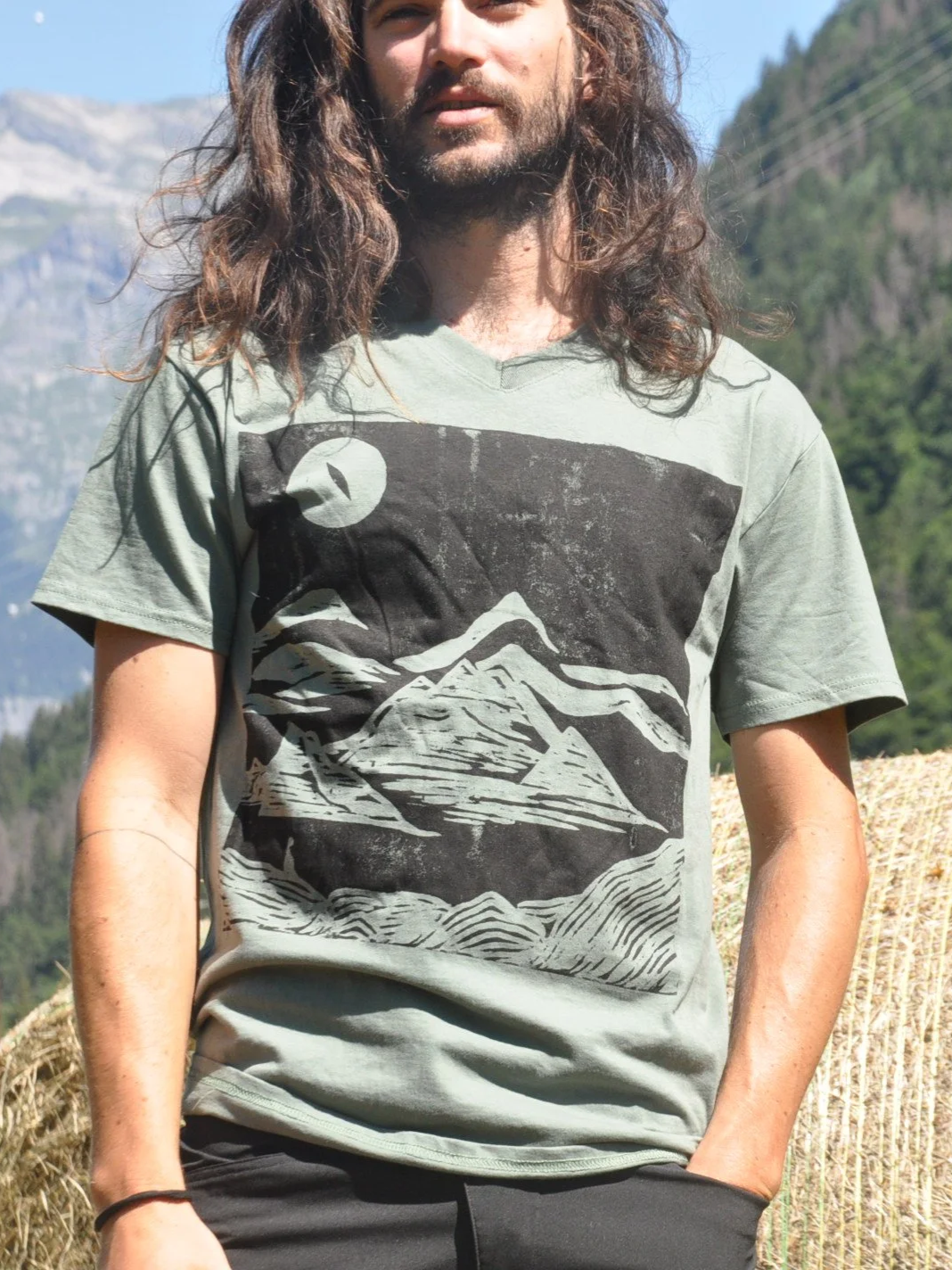 Joven con barba y cabello largo, con camiseta de color verde, en un campo con montañas y árboles de fondo.